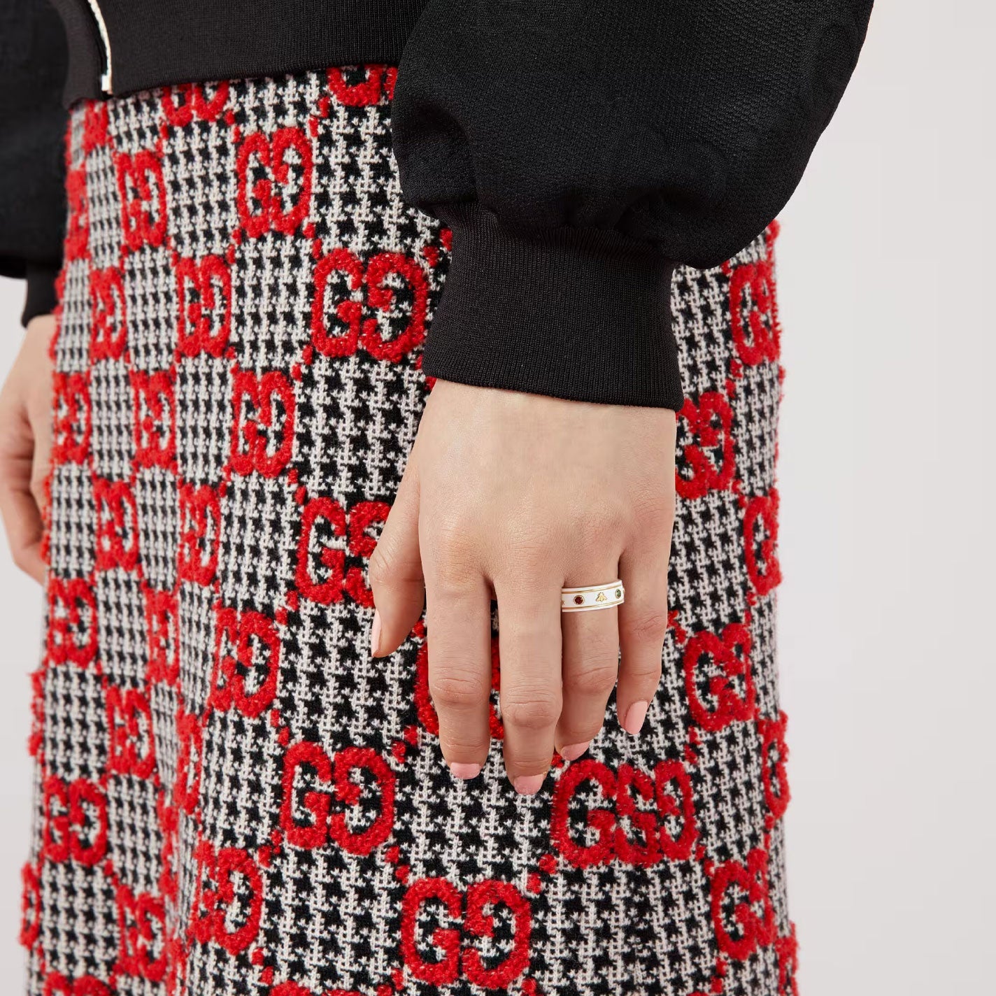 A person wearing a black sweater and a skirt adorned with a red and black checkered pattern featuring the letters "GG" in red, complements their style with a Gucci Icon Ring crafted from 18K Yellow Gold and embellished with White Zirconia and multicolored gemstones on their finger.