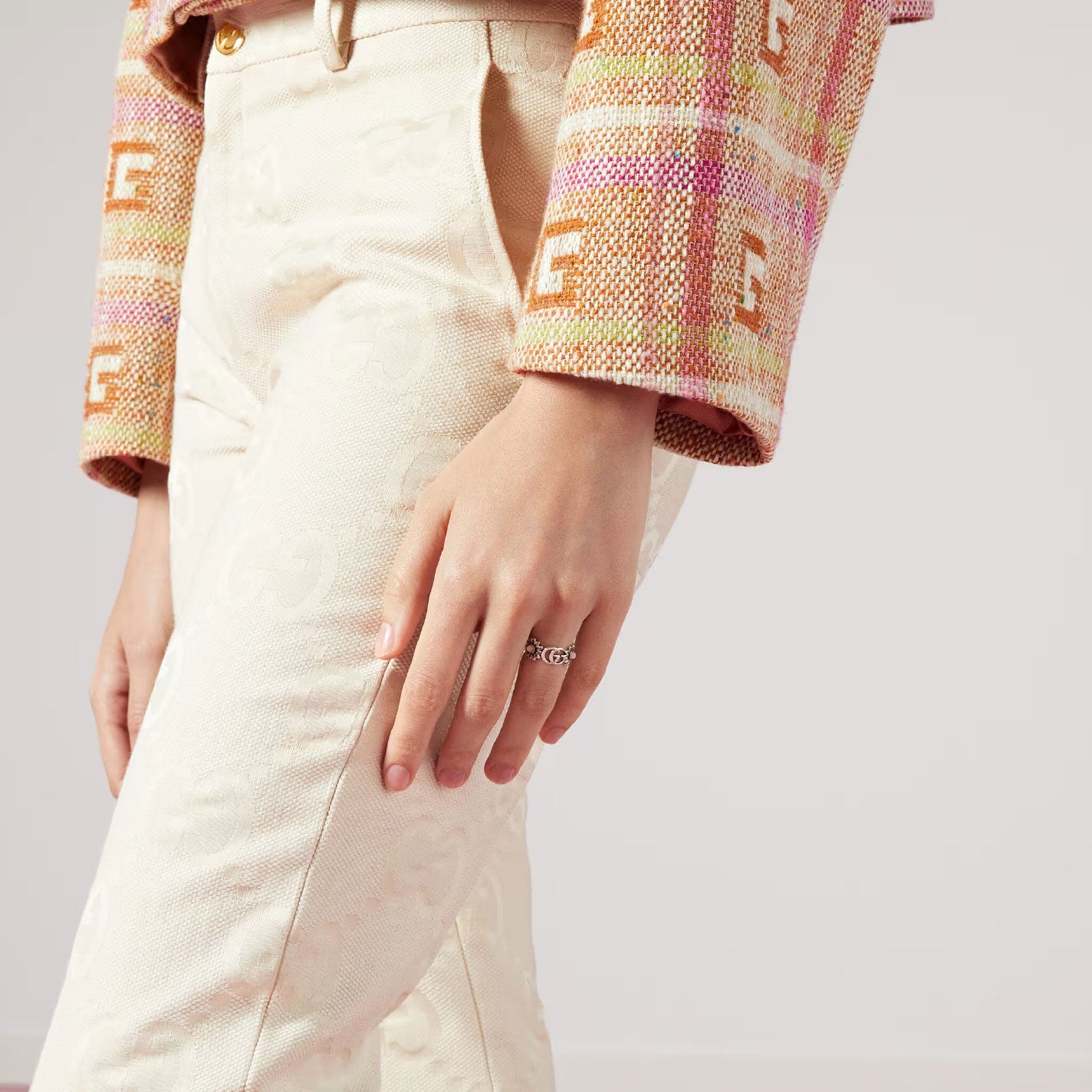 A person wearing light-colored textured pants and a multicolored plaid jacket is seen with their hand, adorned with the Gucci Double G Sterling Silver Ring With Pink Mother Of Pearl from GUCCI Fine Jewellery, resting on their thigh. The jacket features a pattern with letter motifs.