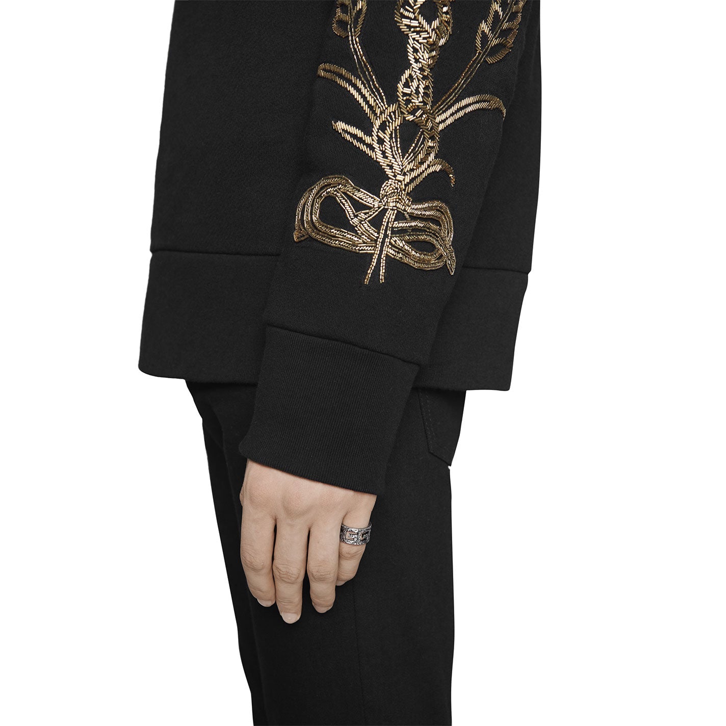 A person dressed in a black ensemble with gold floral embroidery on the sleeve elegantly bends their arm, showcasing the exquisite Gucci G Cube Sterling Silver Ring by GUCCI Fine Jewellery on their index finger, enhancing the look.