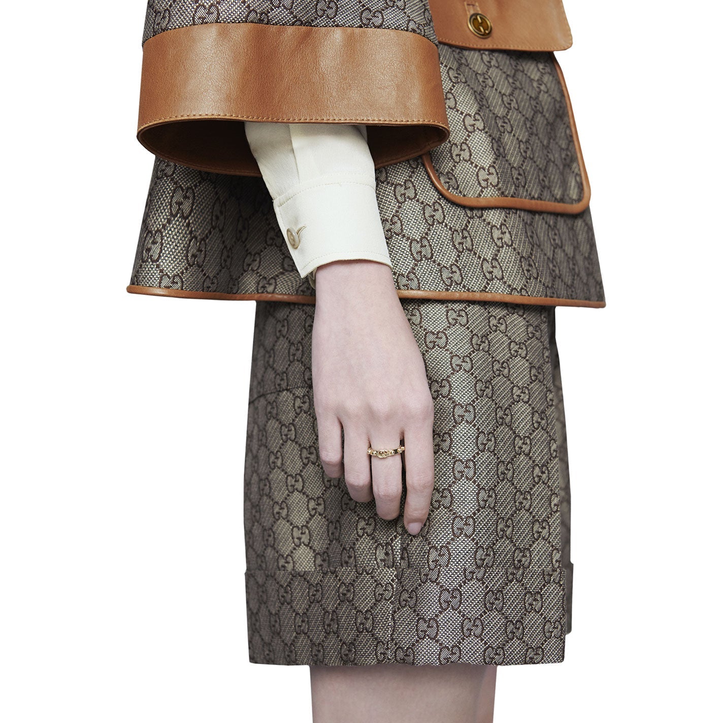 A person dressed in a designer Gucci outfit featuring a monogram pattern and brown leather trim is wearing a matching skirt and jacket. Their hand is adorned with a GUCCI Fine Jewellery Running G 18K Yellow Gold Ring, size 6.75. The background is plain white.