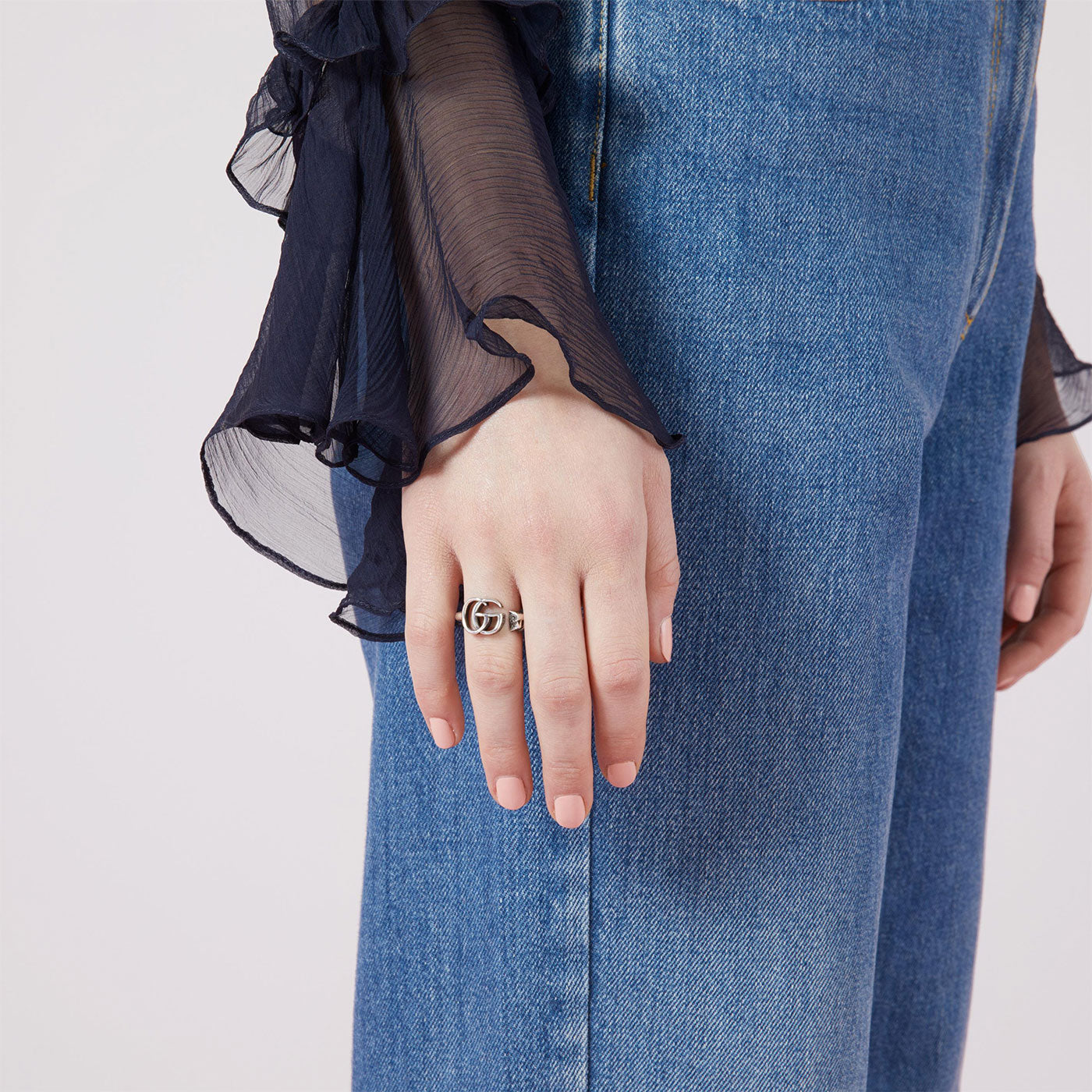 A person in blue jeans and a sheer black blouse is depicted. Their left hand, featuring the Gucci GG Marmont Sterling Silver Key Ring from GUCCI Fine Jewellery, rests against their leg. The emphasis is on the hand and the key ring.
