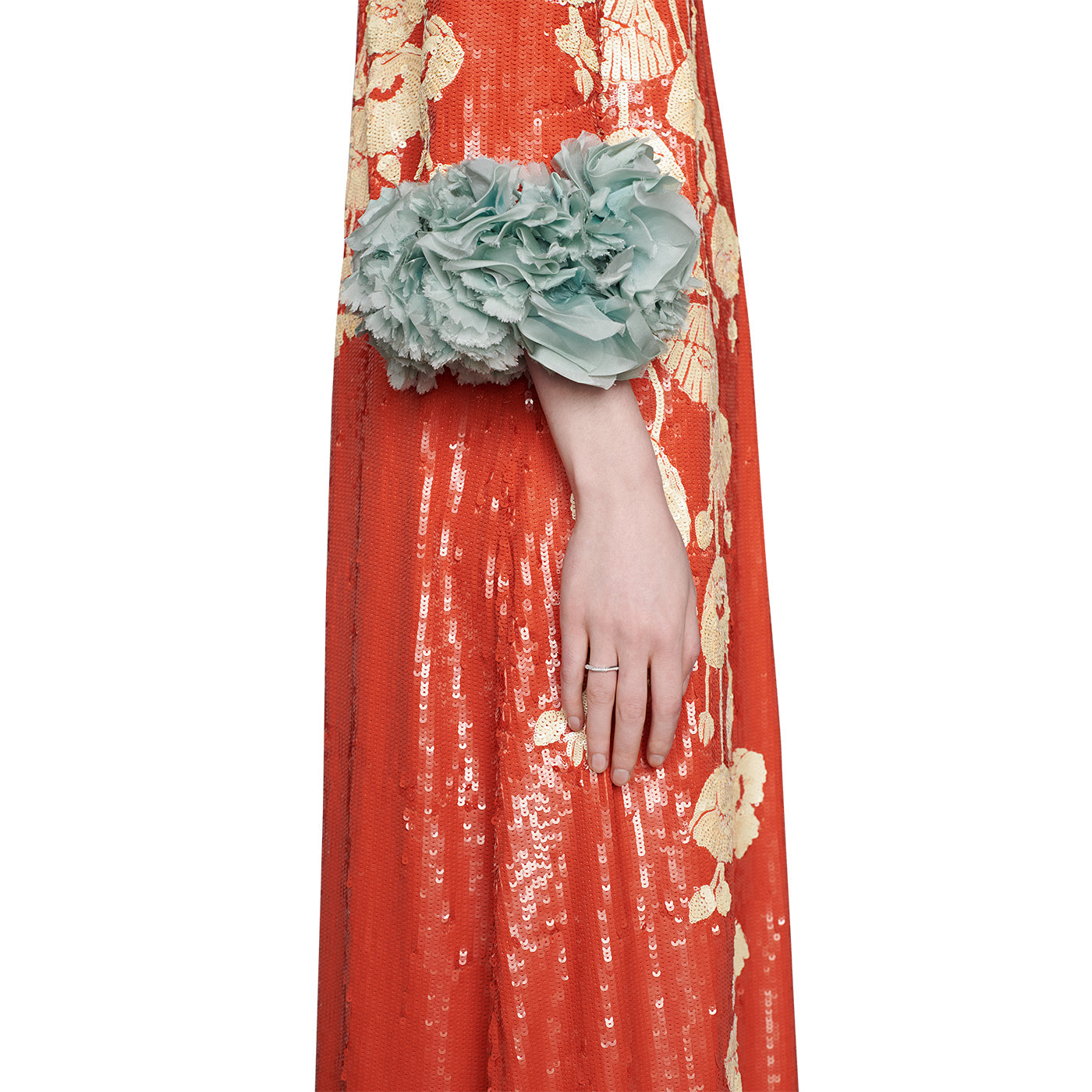 A person wearing a vibrant red and gold embroidered gown adorned with sequins casually rests their hand on their side, showcasing the elegant GUCCI Fine Jewellery Link to Love 18K White Gold Diamond Ring with a stackable mechanism. The sleeve features a large cluster of light blue flowers at the elbow.