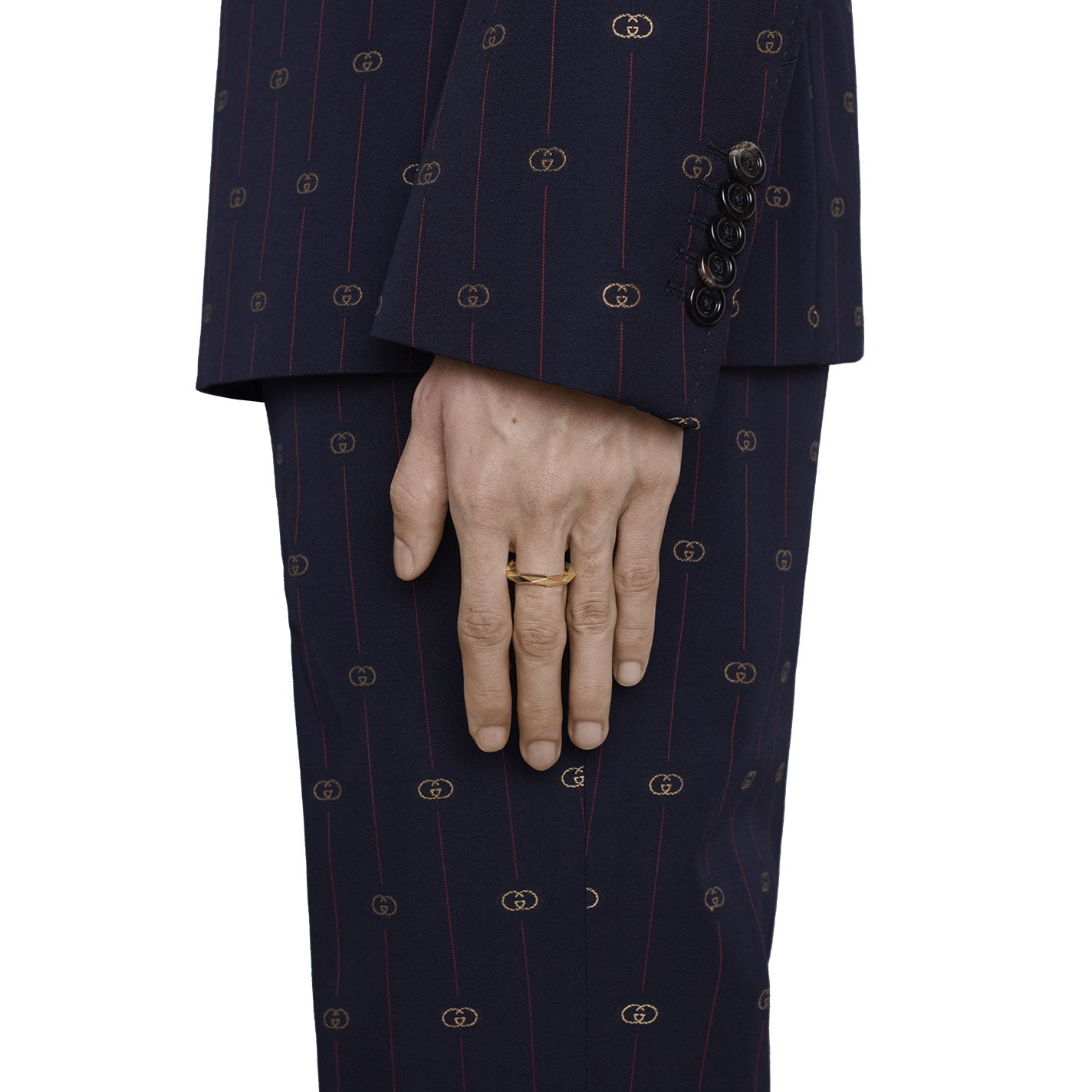 A person wearing a dark, patterned suit jacket and matching pants has a Gucci Link to Love Studded 18K Yellow Gold Ring on their finger from GUCCI Fine Jewellery. The suit features discreet circular motifs, and their hand rests casually by their side.