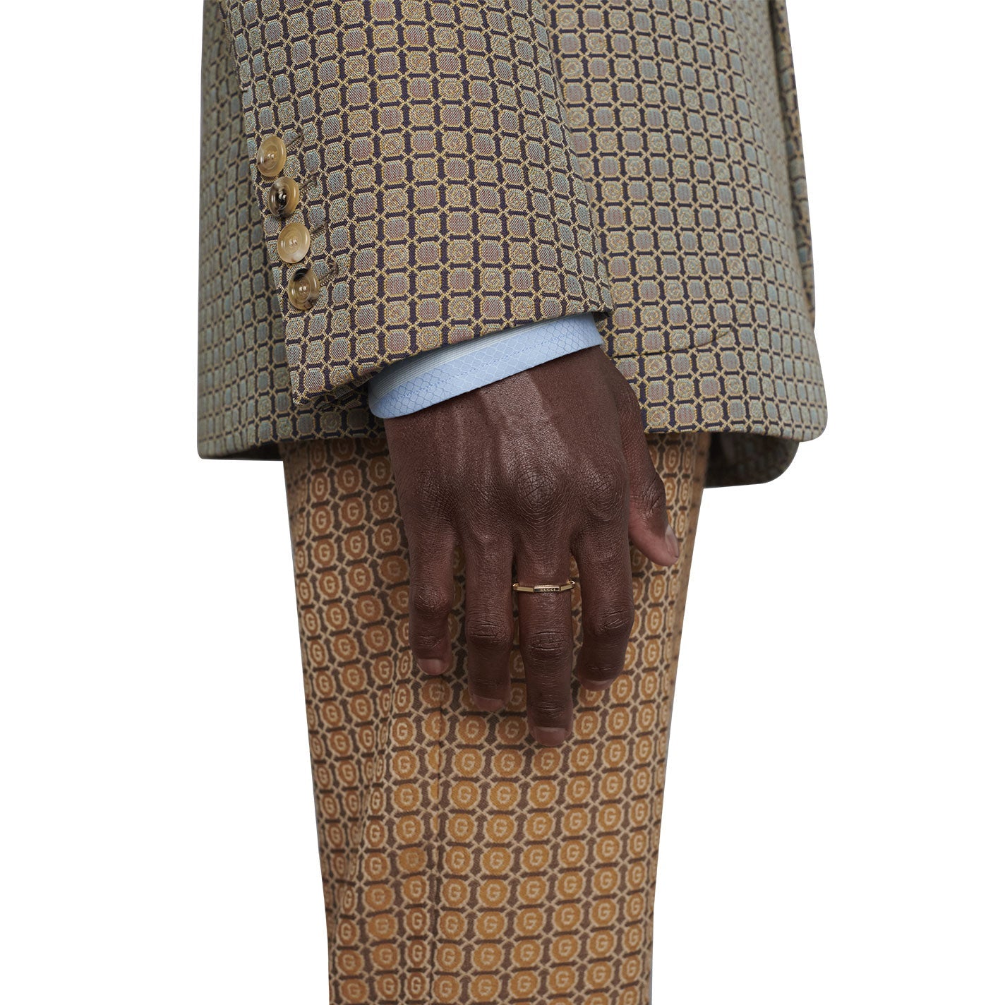 Wearing a beige patterned suit with a light blue shirt underneath, their hand, adorned with the elegant Gucci Link to Love Mirrored 18K Yellow Gold Ring from GUCCI Fine Jewellery, rests relaxed by their side against matching patterned trousers.