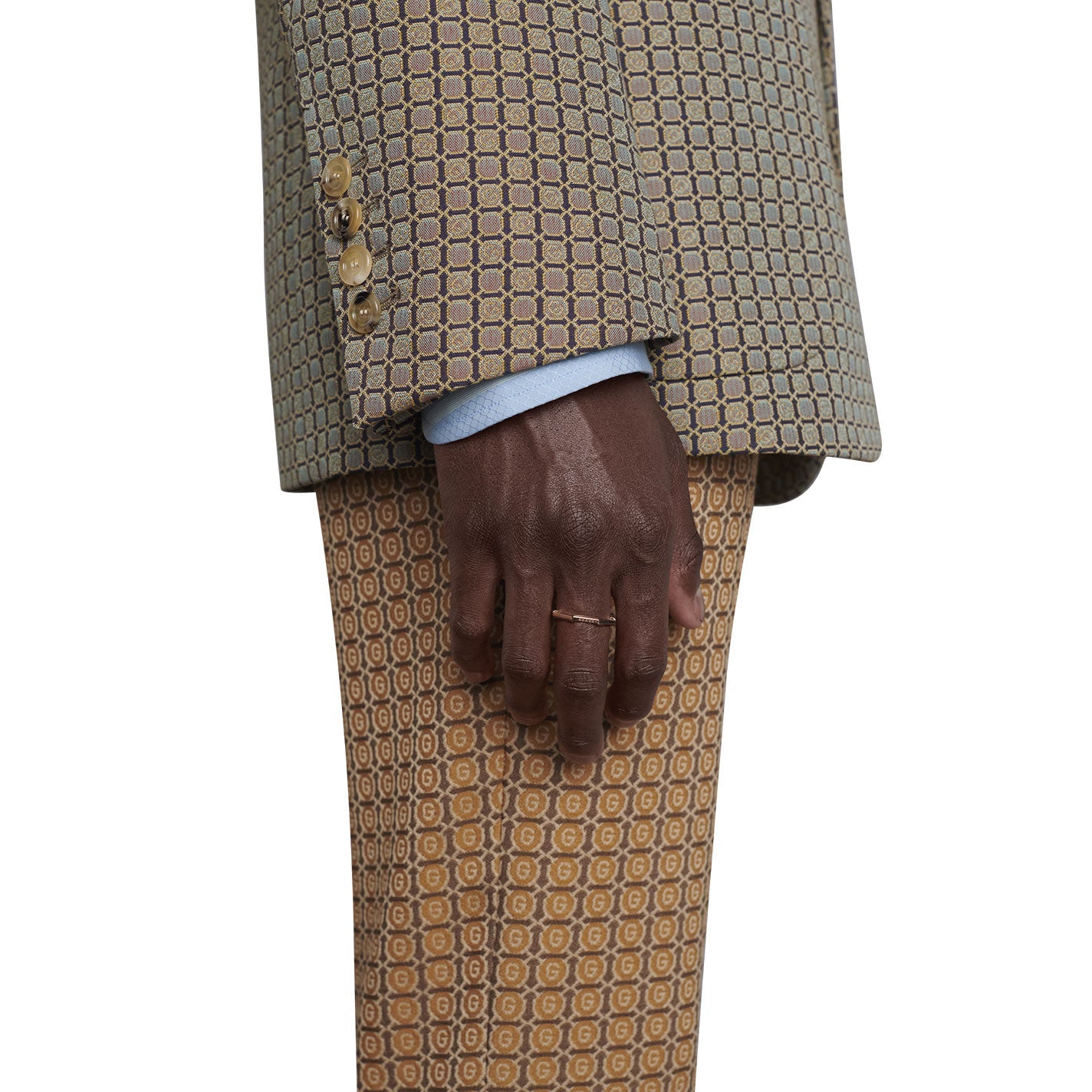 A person with dark skin wearing a patterned beige suit stands with their hand resting on their leg. The suit boasts a geometric design, and the sleeve is adorned with beige buttons. Elegantly displayed on their finger is a Gucci Link to Love Mirrored 18K Rose Gold Ring from GUCCI Fine Jewellery.