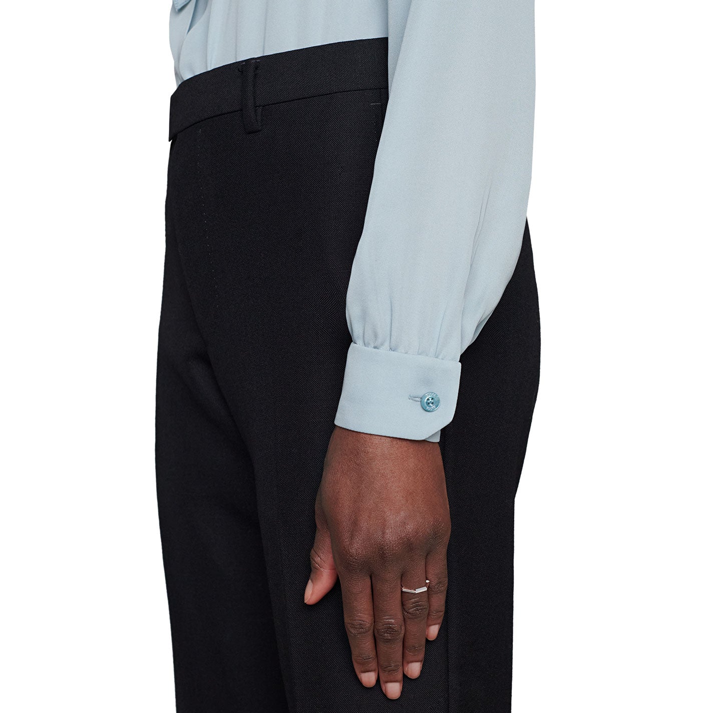 A person is dressed in a light blue long-sleeve blouse with buttoned cuffs and black pants. Their hand, adorned with a Gucci Link to Love Mirrored 18K White Gold Ring from GUCCI Fine Jewellery, rests at their side. The image captures a partial side view against a white background.