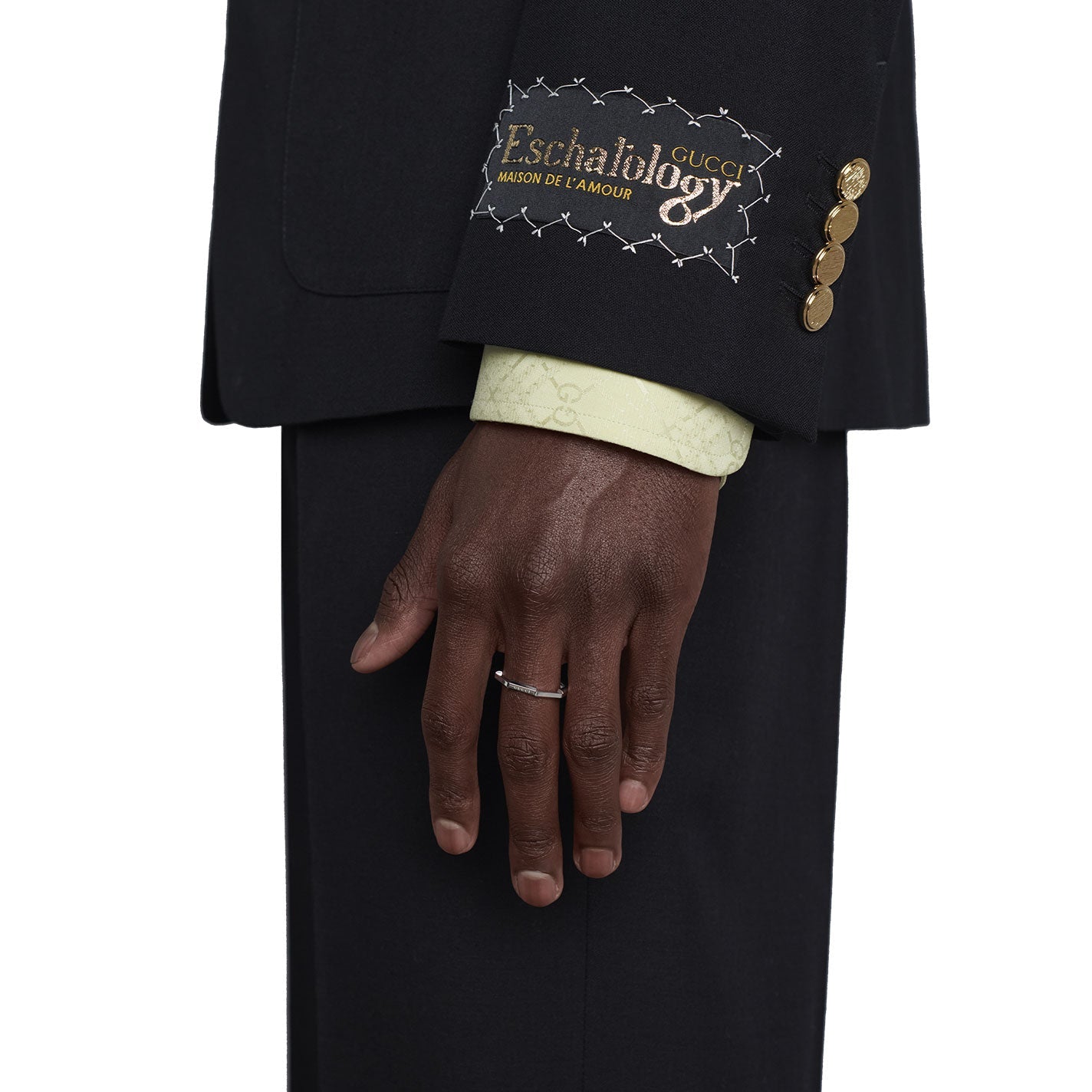 A close-up of a person wearing a dark suit with gold buttons on the sleeve and decorative stitching that reads "Maison de l'Amour." The person's hand is visible, adorned with a Gucci Link to Love Mirrored 18K White Gold Ring from GUCCI Fine Jewellery, over a light patterned shirt cuff.
