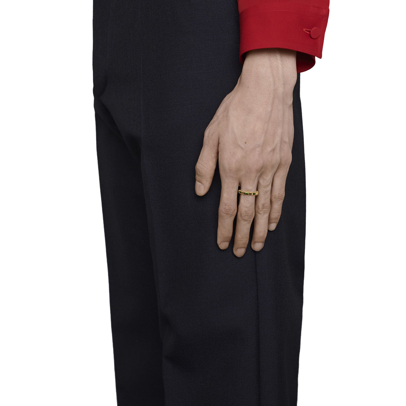 A person wearing a red shirt has their right hand resting on dark pants, showcasing a sleek Gucci Link to Love Tourmaline 18K yellow gold ring from GUCCI Fine Jewellery on the ring finger. The background is white.