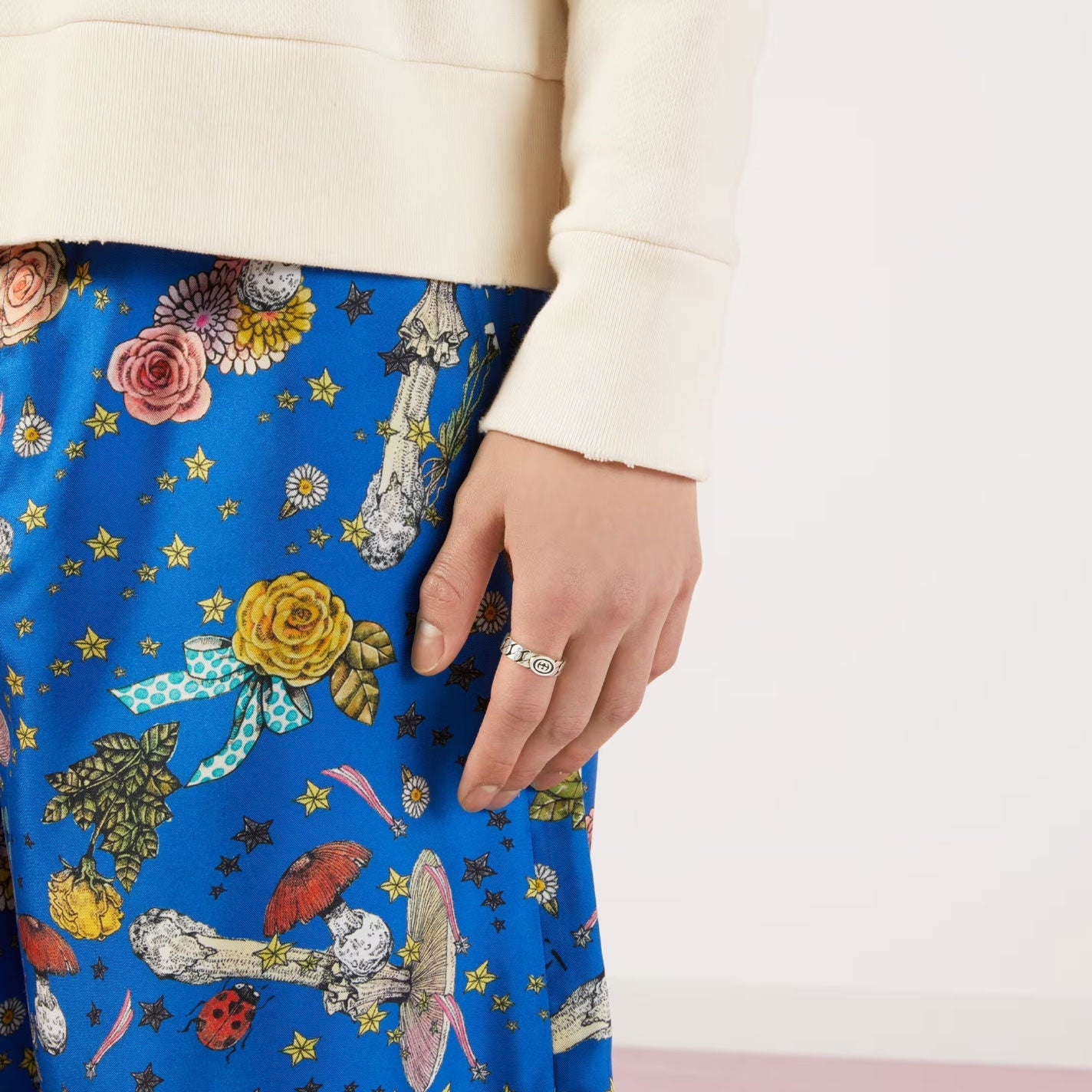 A person in a cream top and vibrant pants decorated with floral and celestial designs, displaying an enameled Interlocking G. Their hand, highlighted by a Gucci Interlocking G Wide Sterling Silver Ring from the GUCCI Fine Jewellery collection, gently rests against a plain, light-colored wall.