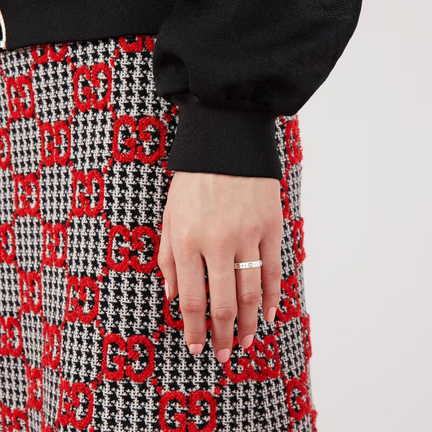 A person wearing a black long-sleeve top and a checkered skirt with red and black patterns rests their hand naturally against their leg, showcasing two rings: the Gucci Icon Interlocking G White Zirconia and 18K Yellow Gold Ring from GUCCI Fine Jewellery. The repeating logo pattern on the skirt in red reflects Gucci's iconic style.