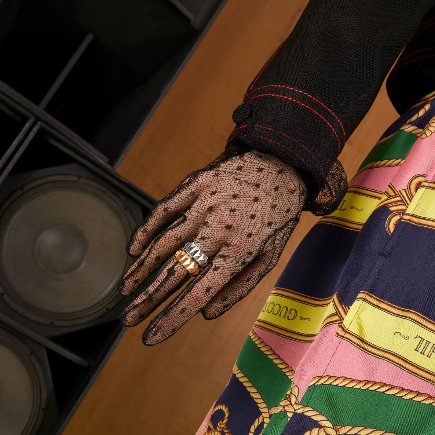 A hand adorned with black mesh gloves displays a stackable collection of gold rings, highlighted by the elegant Gucci Link to Love Studded 18K Yellow Gold Ring from GUCCI Fine Jewellery, set against a backdrop of sound equipment and a vibrant patterned dress with ropes and chains.