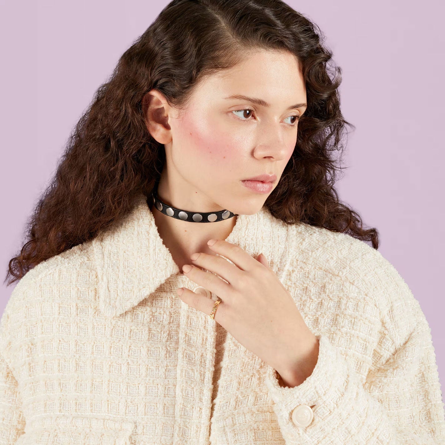 A person with long, curly brown hair is seen wearing a textured cream jacket and a black choker adorned with round silver accents, glancing sideways against a pale purple background. A subtle nod to the Gucci Icon Star 18K Yellow Gold Ring from GUCCI Fine Jewellery adds an elegant touch.