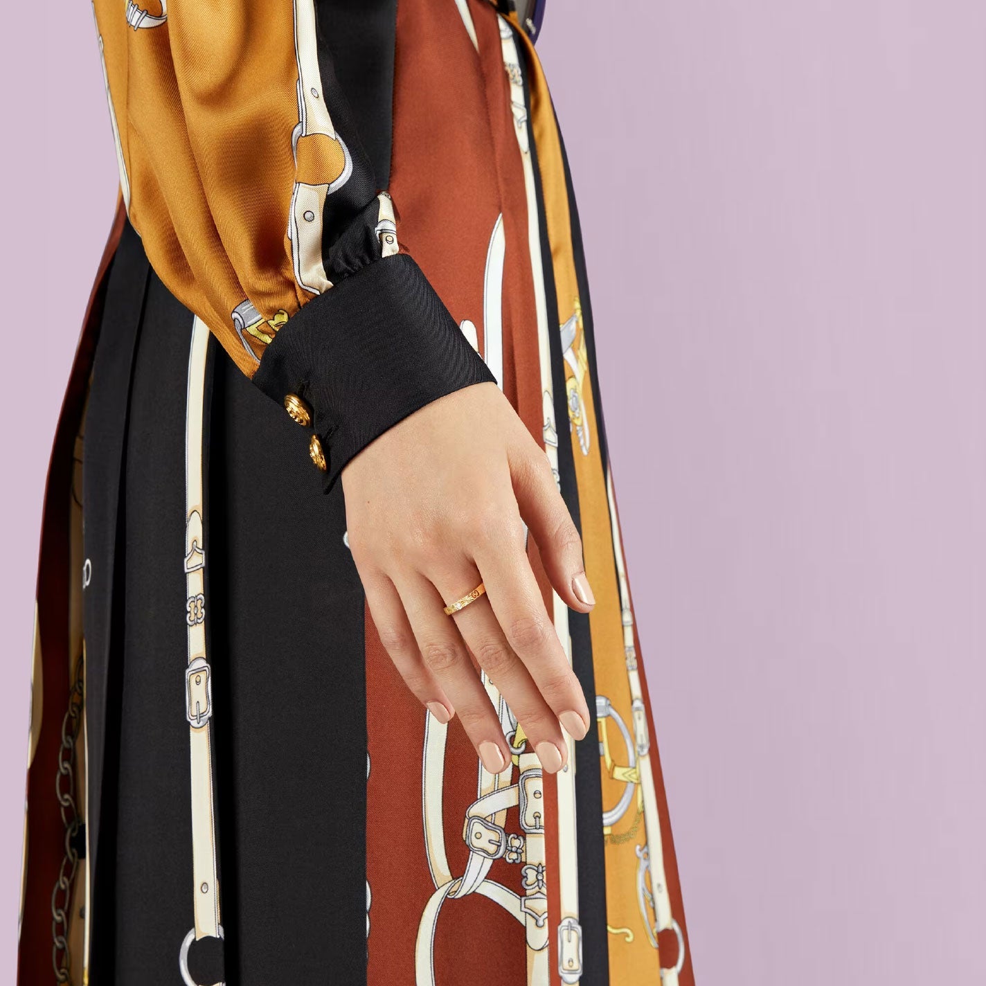 A close-up of a woman's hand adorned with the Gucci Icon Heart 18K Rosé Gold Diamond Ring from GUCCI Fine Jewellery, resting against her colorful dress with patterns of belts and chains. Her attire showcases black, orange, and golden hues amidst the elegant glow. The background is a soft lavender color, enhancing the scene's grace.