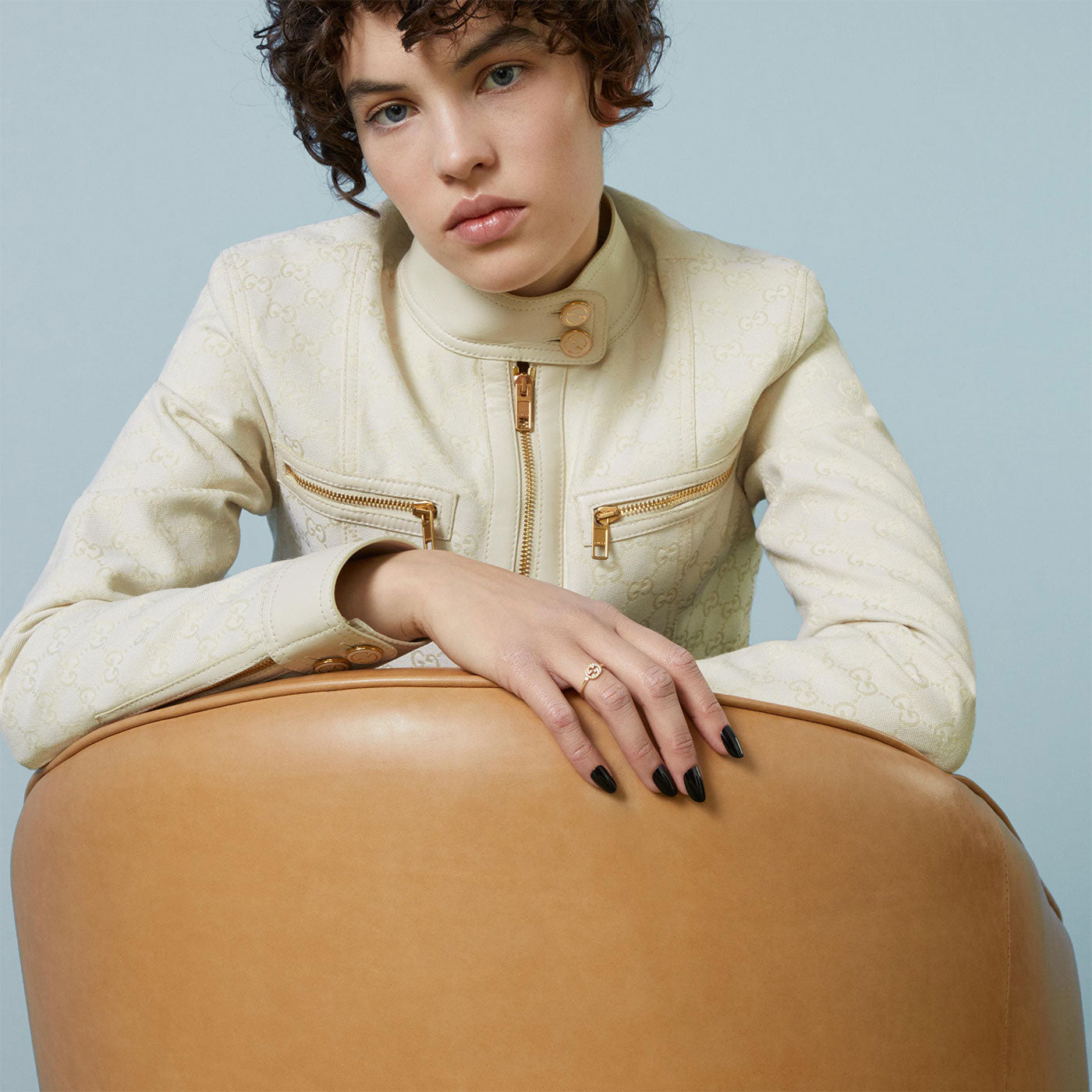 A person with short curly hair leans on a tan leather chair, dressed in a cream-colored jacket featuring a high collar and zippers. They sport black-painted nails and wear an expression of relaxation, enhanced by the elegant Gucci Interlocking G 18K Yellow Gold Diamond Ring. The muted blue background adds to the sophisticated allure of this GUCCI Fine Jewellery piece.
