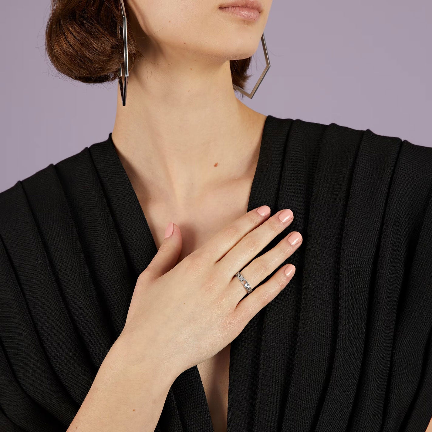 A person wearing large geometric earrings and a black pleated top rests a hand adorned with the Gucci Icon Heart 18K White Gold Ring on their chest. The subtle sheen of the GUCCI Fine Jewellery piece adds an elegant touch against the soft purple background, enhancing their look.