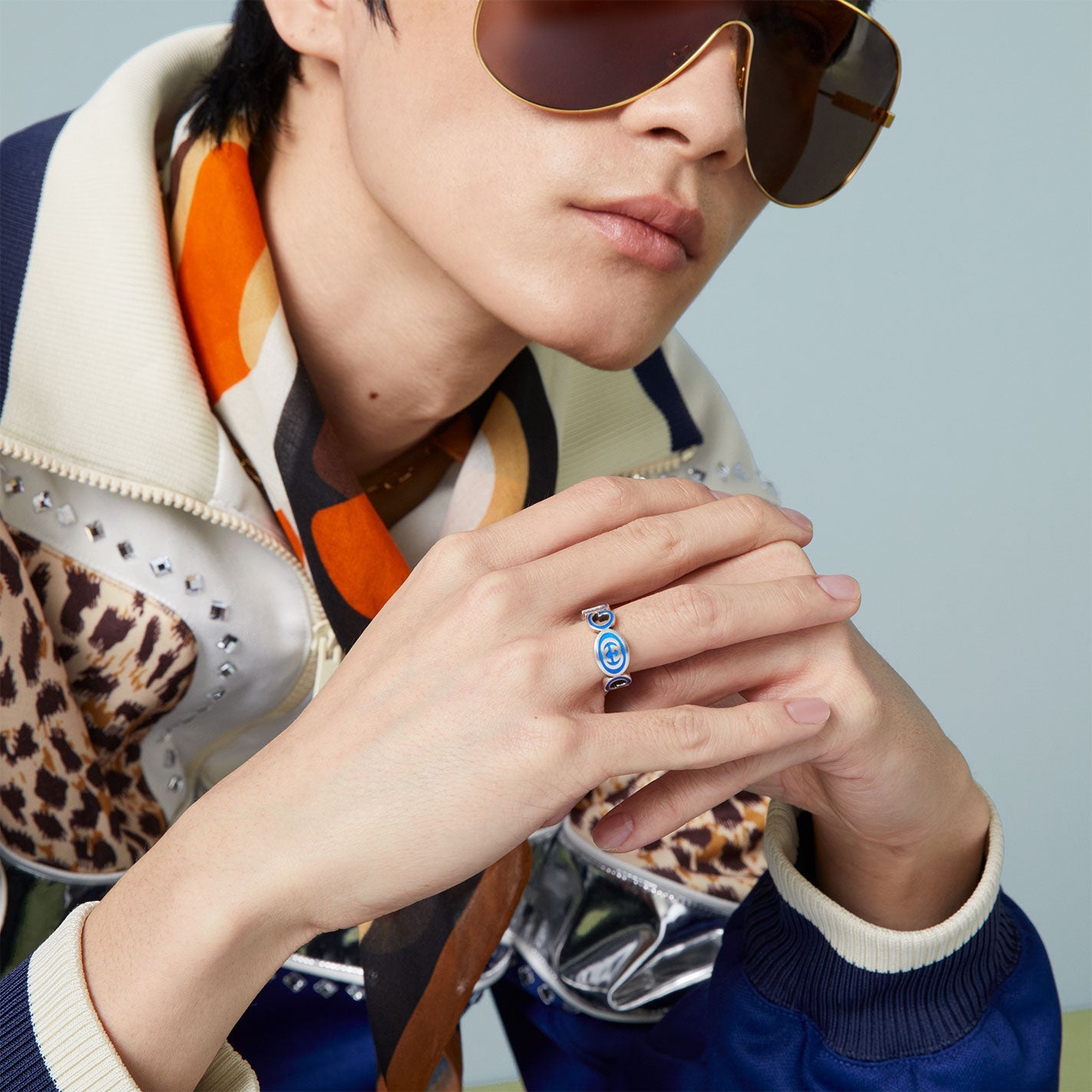A person wearing large sunglasses and a colorful scarf rests their chin on clasped hands, adorned with a Gucci Interlocking G Enamel Sterling Silver Ring from GUCCI Fine Jewellery. Their outfit includes a jacket with leopard print and silver studs, perfectly capturing an edgy yet chic style.