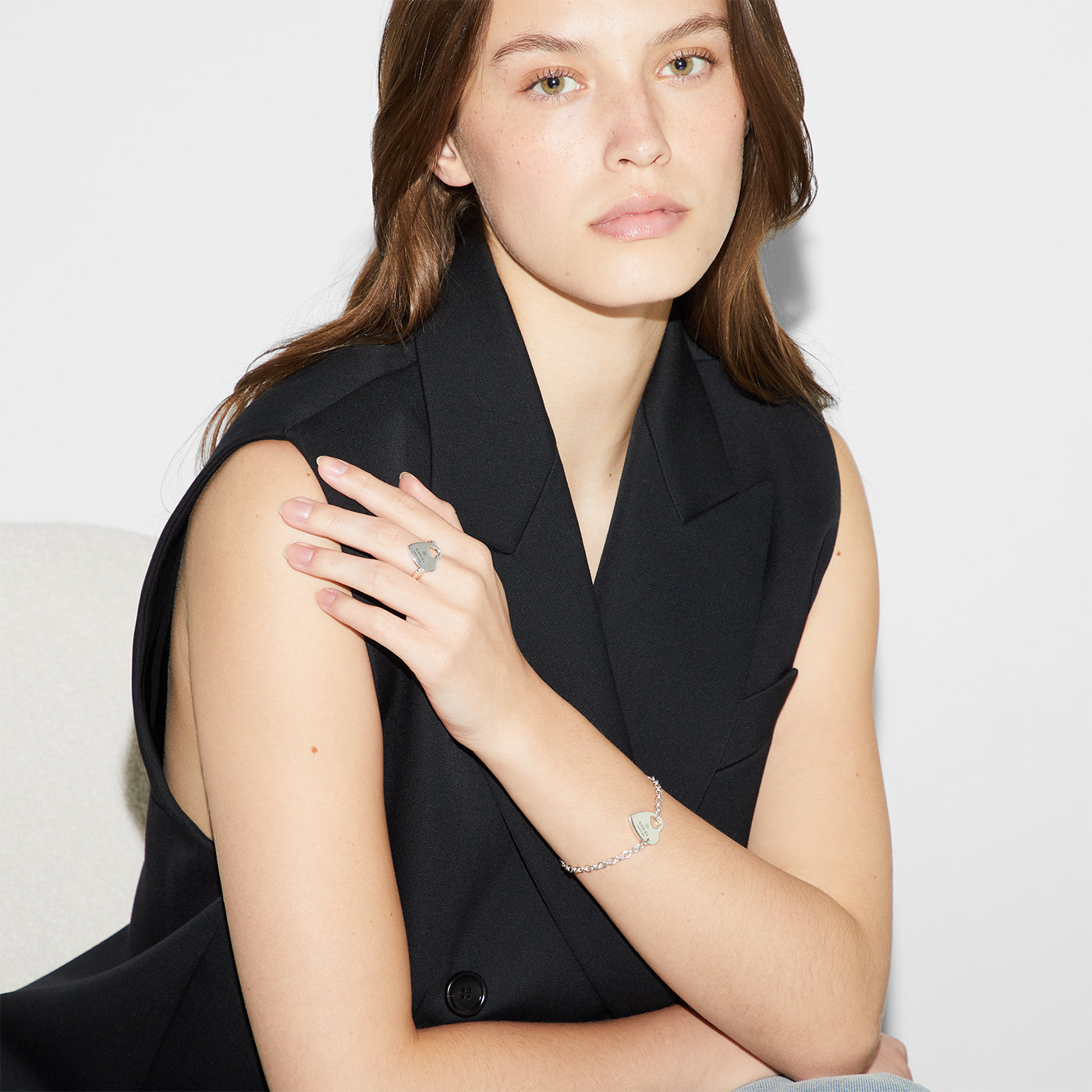 A woman with long brown hair sits in a black sleeveless blazer, displaying the Gucci Trademark Sterling Silver Heart Ring accompanied by a bracelet featuring matching designs. Her neutral expression captures the timeless elegance of GUCCI Fine Jewellery's Pre-Fall 2024 collection against a plain white backdrop.