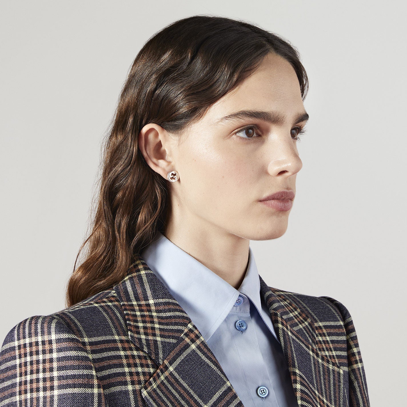 A person with long, wavy brown hair is wearing a plaid blazer over a light blue shirt. They have a neutral expression and small, round Gucci Interlocking G Sterling Silver Stud Earrings from GUCCI Fine Jewellery. The background is plain and light-colored.