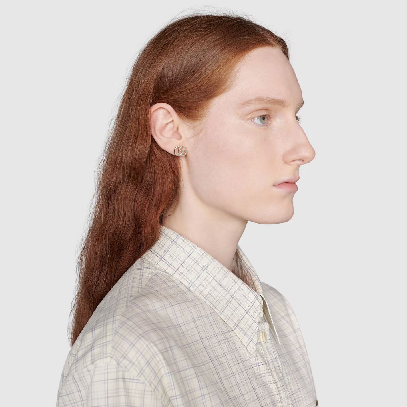 A person with long auburn hair is wearing a plaid shirt and a GUCCI Fine Jewellery Lionhead 18K Yellow Gold Chromodiopside and Diamond Stud Earring. They are shown in profile against a plain gray background.