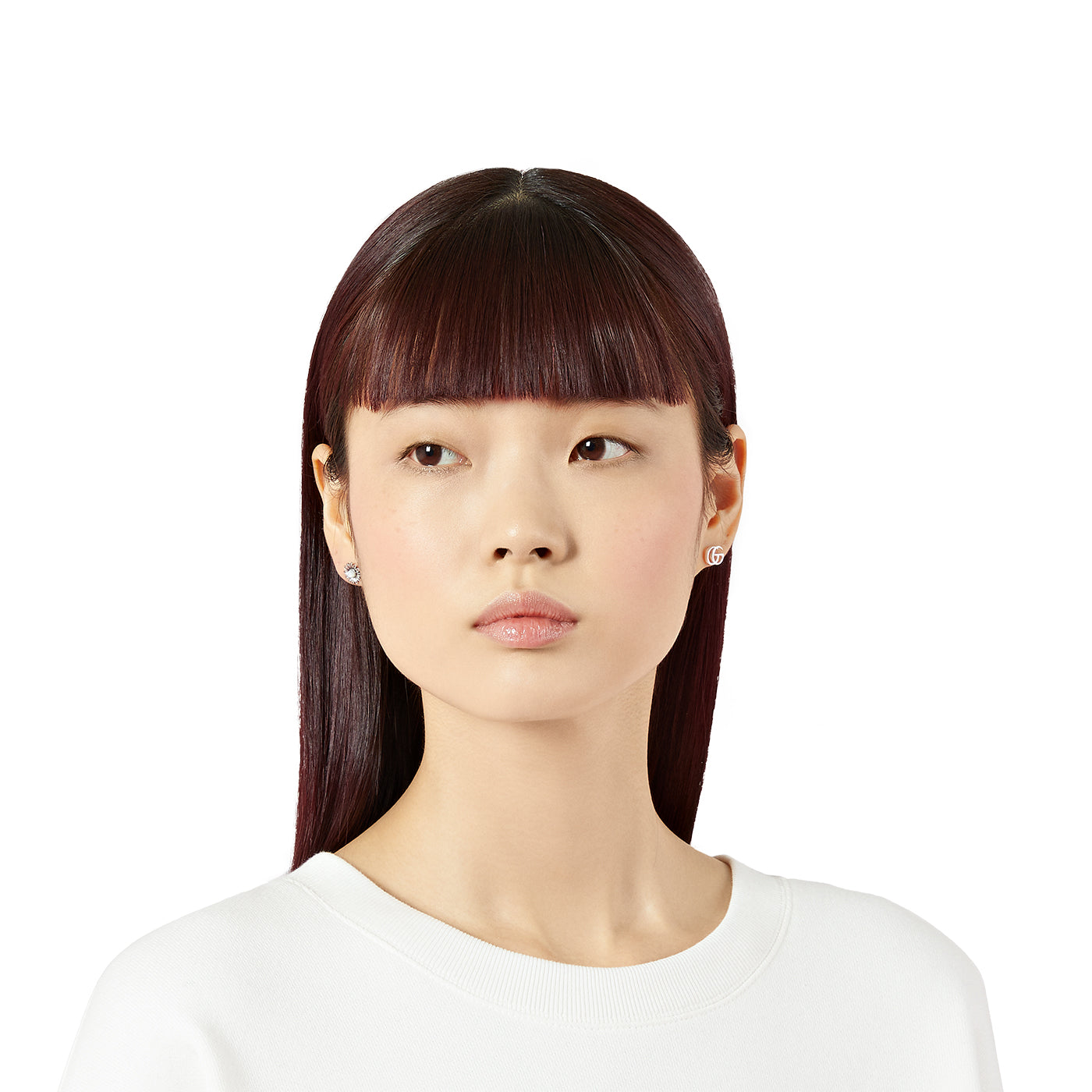 A person with straight, dark hair and bangs is wearing a white top accented by a GUCCI Fine Jewellery stud earring featuring the Double G sterling silver design with a mother-of-pearl flower. They are looking to the side with a neutral expression against a plain white background.