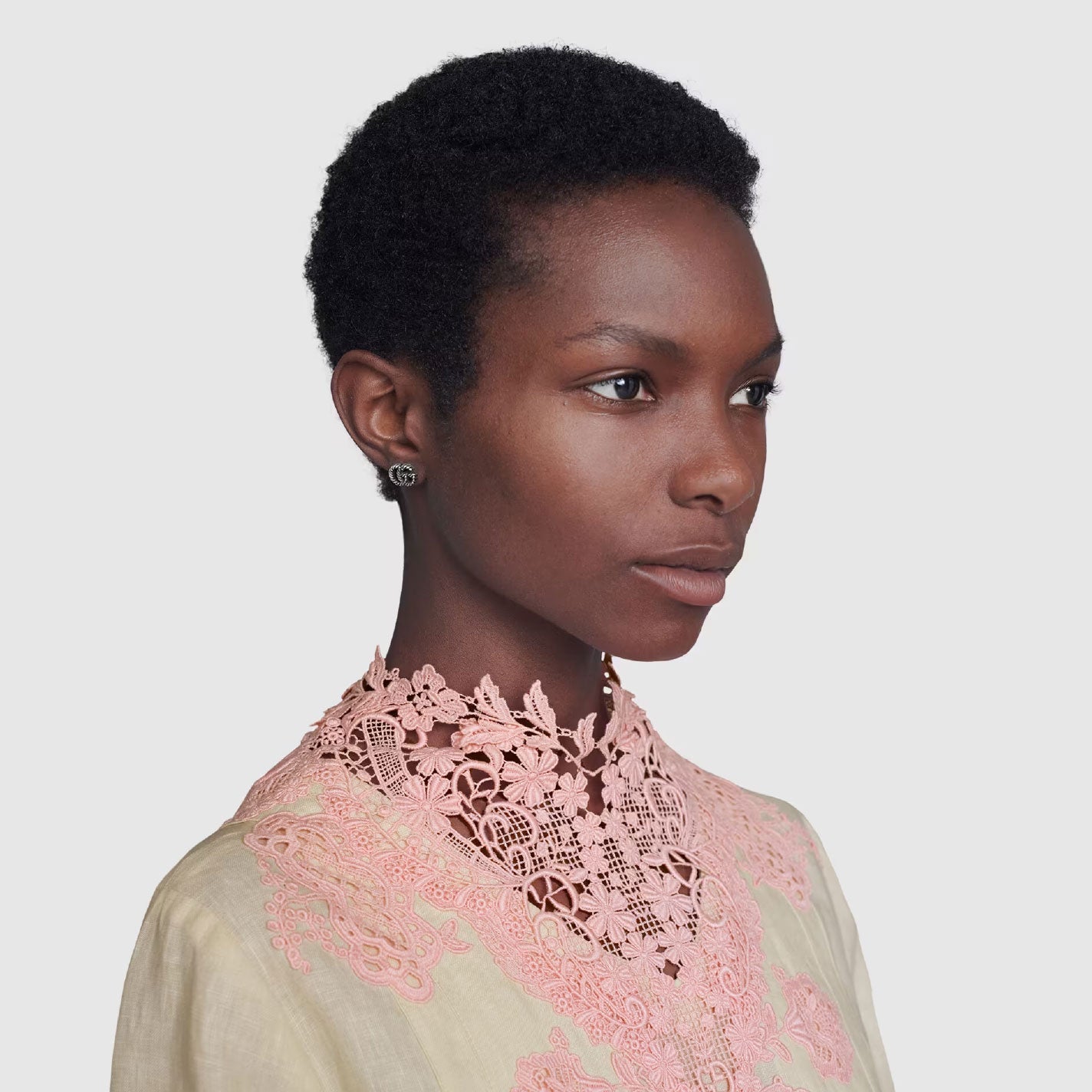 A person with short curly hair wears a delicate light pink lace top against a plain background, gazing to the side. They have Gucci Double G Sterling Silver Stud Earrings by GUCCI Fine Jewellery and a composed expression.