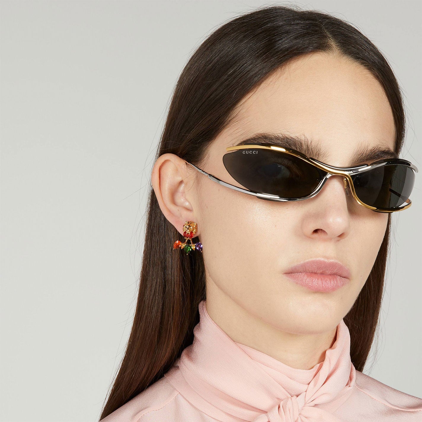 A person with straight brown hair wears sleek black sunglasses and a vibrant, dangling Gucci Lionhead 18K Yellow Gold Diamond and Multi Colour Gemstone Single Stud Earring by GUCCI Fine Jewellery. They are dressed in a light pink, high-collared top against a plain background.