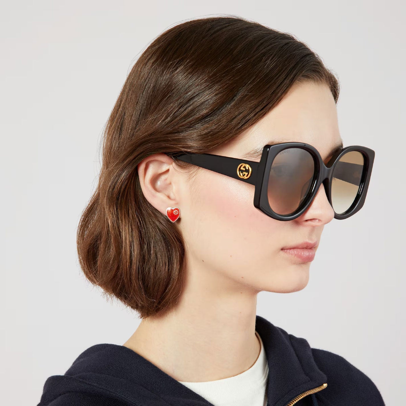 A person with short brown hair wears oversized black sunglasses adorned with a gold logo. They sport Gucci Interlocking G sterling silver stud earrings with red enamel from GUCCI Fine Jewellery, paired with a white shirt and dark top, standing against a neutral background.