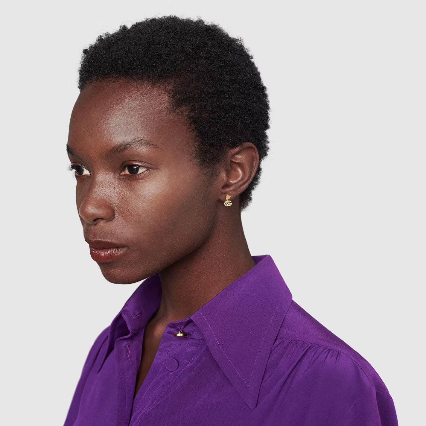 A person with short black hair is wearing a vibrant purple shirt and small Gucci Running G 18K Yellow Gold Stud Earrings by GUCCI Fine Jewellery. The background is a plain, light gray color. The person looks pensive and slightly faces to the right.