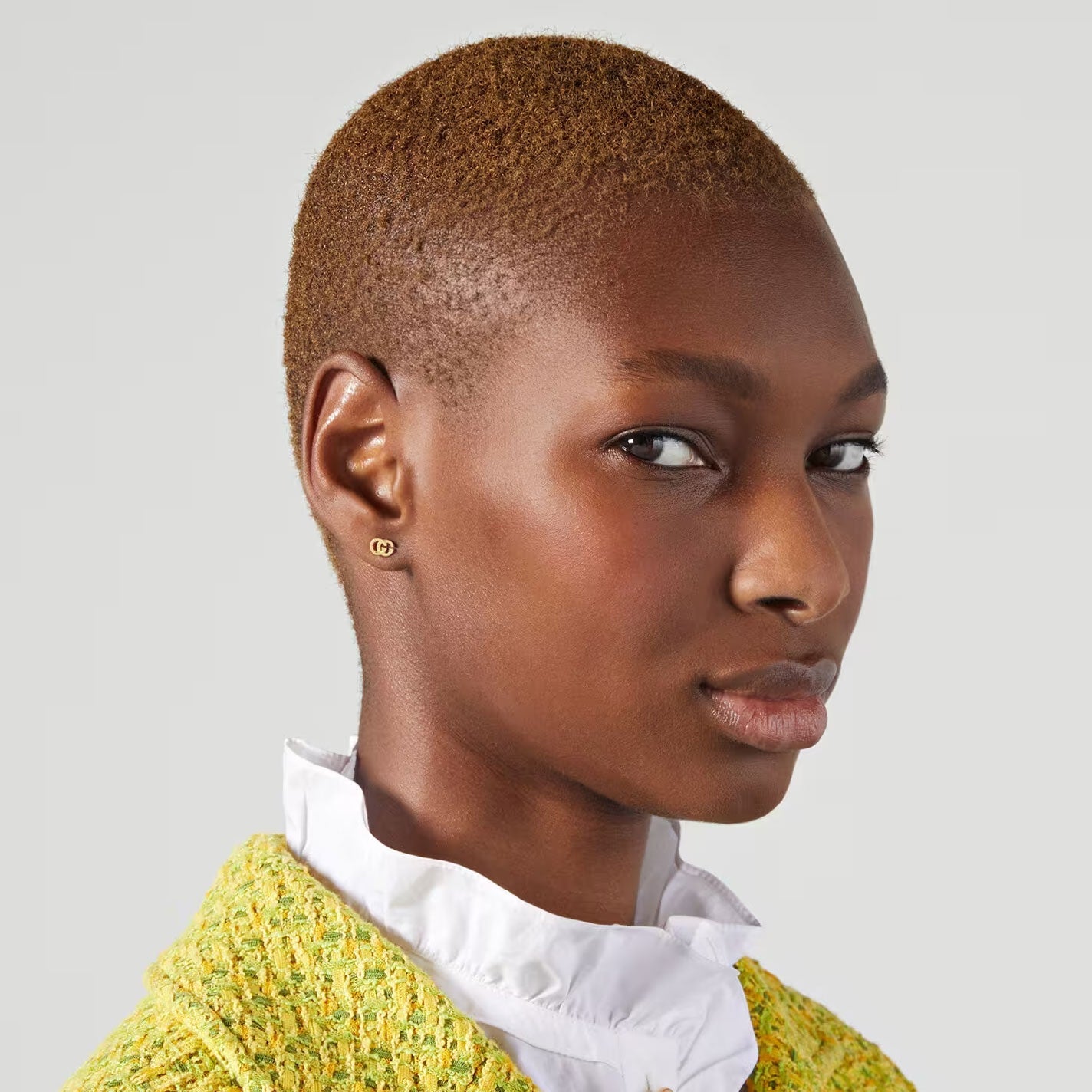 A person with short hair wears a textured yellow jacket over a white shirt, accessorized with Gucci Running G 18K Yellow Gold Stud Earrings from GUCCI Fine Jewellery, exuding elegance. They have a calm expression and gaze slightly to the side, against a plain gray background.