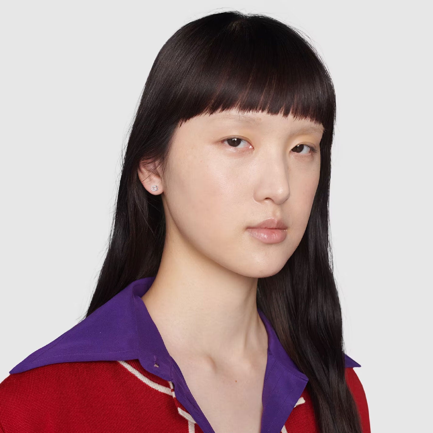 A person with long black hair and blunt bangs, dressed in a red sweater featuring a purple collar, has a neutral expression as they glance slightly towards the camera. The backdrop is a plain light gray, embodying the minimalist aesthetic frequently associated with GUCCI Fine Jewellery, such as the Gucci Running G 18K White Gold Stud Earrings.