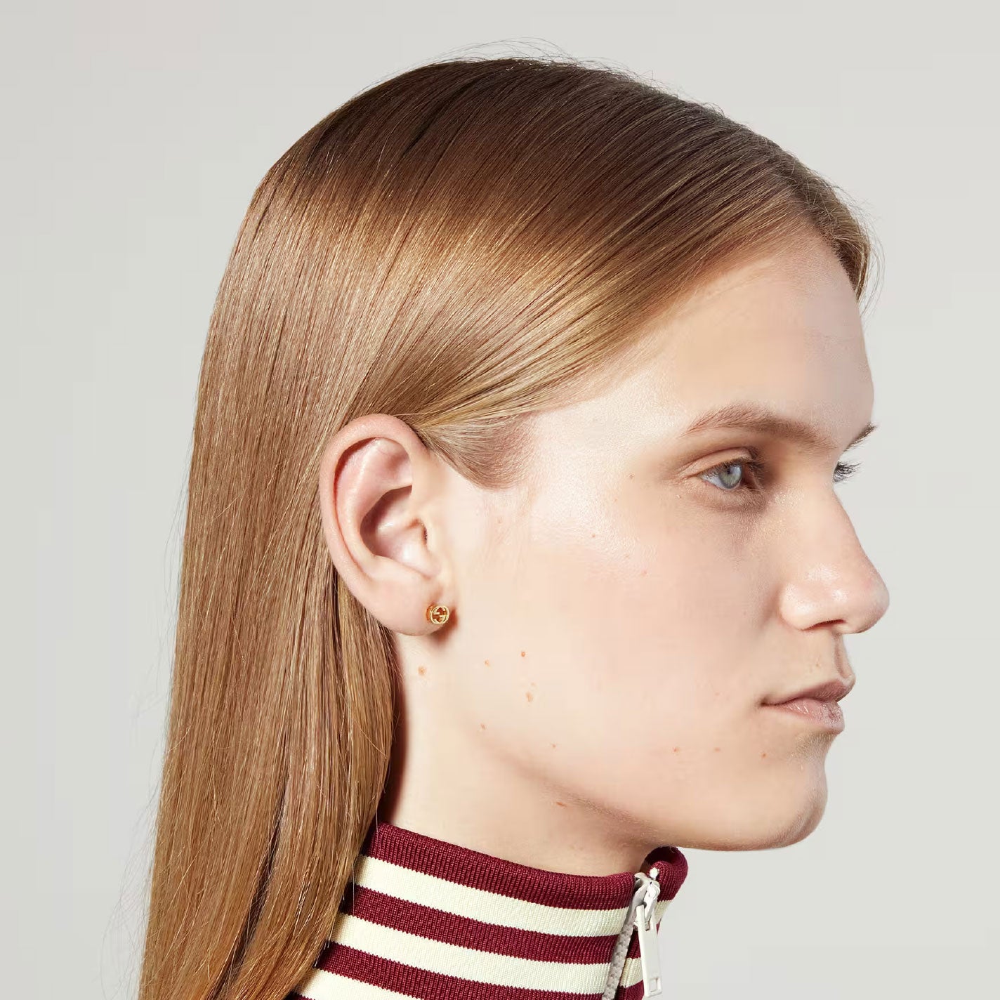 Side profile of a woman with straight, light brown hair wearing a striped, high-necked top. Her small Gucci Interlocking G 18K Yellow Gold Stud Earrings by GUCCI Fine Jewellery glint subtly as she maintains a neutral expression against a plain background.