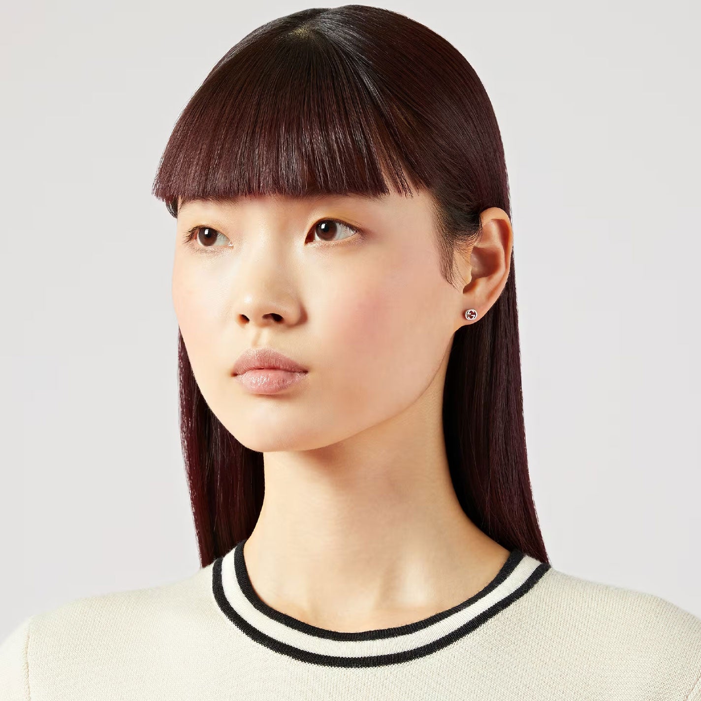 A person with straight, dark brown hair and bangs is wearing a white top featuring a black-striped collar. They have an impassive expression and are turned slightly to the left against a plain backdrop while wearing the Interlocking G 18K White Gold Stud Earrings from GUCCI Fine Jewellery.
