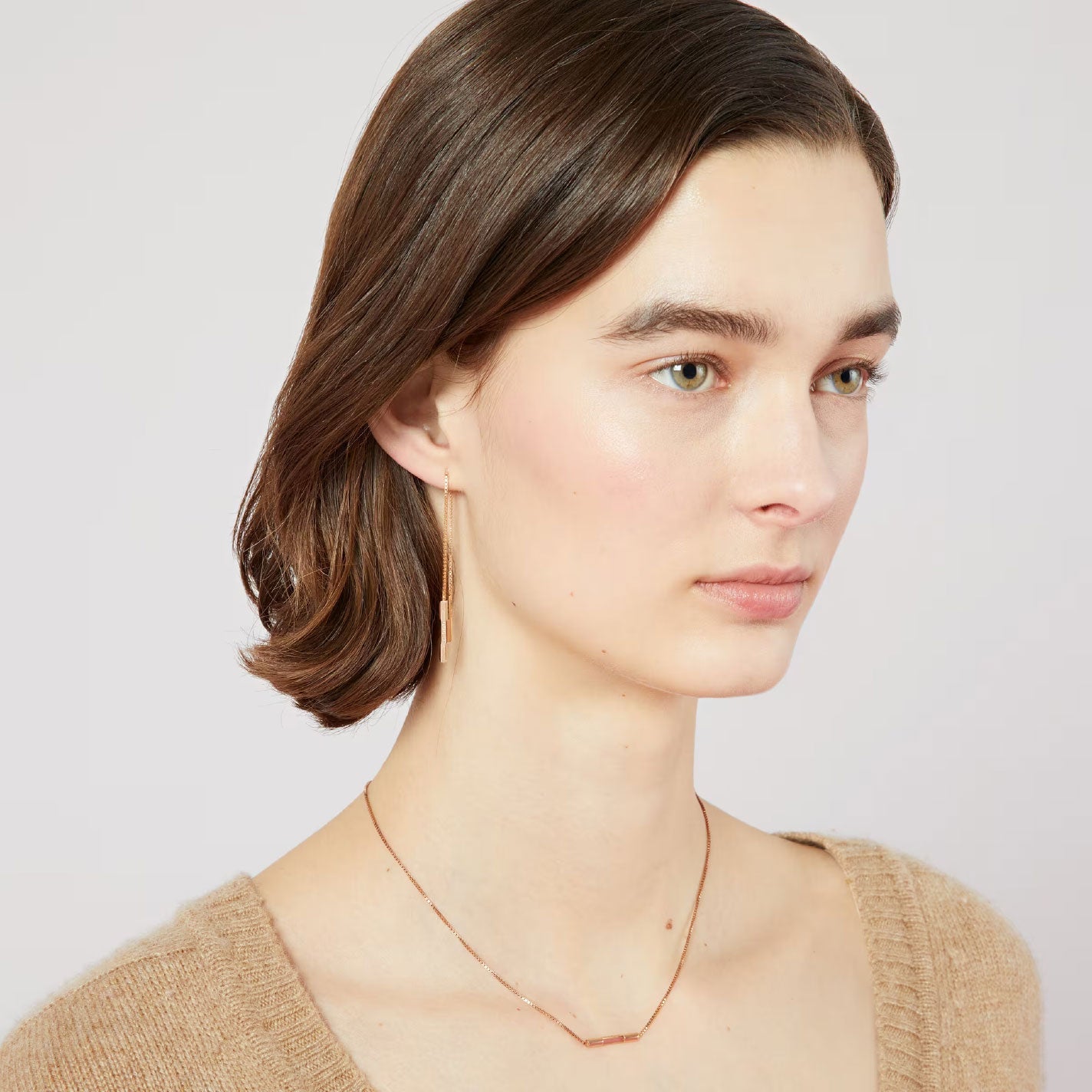 A person with short brown hair is wearing a brown sweater and elegant GUCCI Fine Jewellery. Their thin 18K rose gold necklace pairs beautifully with the Gucci Link To Love 18K Rose Gold Chain Drop Earrings with "Gucci" Bar as they gaze to the side against a plain background.