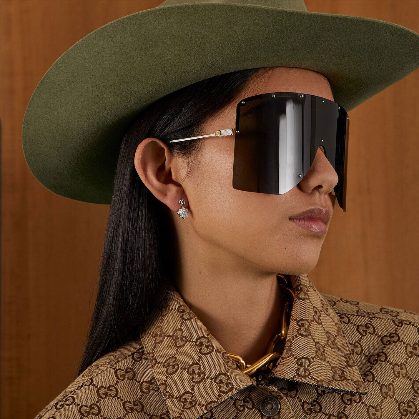 A person clad in oversized dark sunglasses and a green wide-brimmed hat dons a tan patterned Gucci Flora jacket. They accessorize with Gucci Flora Double G 18K White Gold Diamond Stud Earrings from GUCCI Fine Jewellery and wear a chunky 18K white gold necklace, all set against a wooden background.