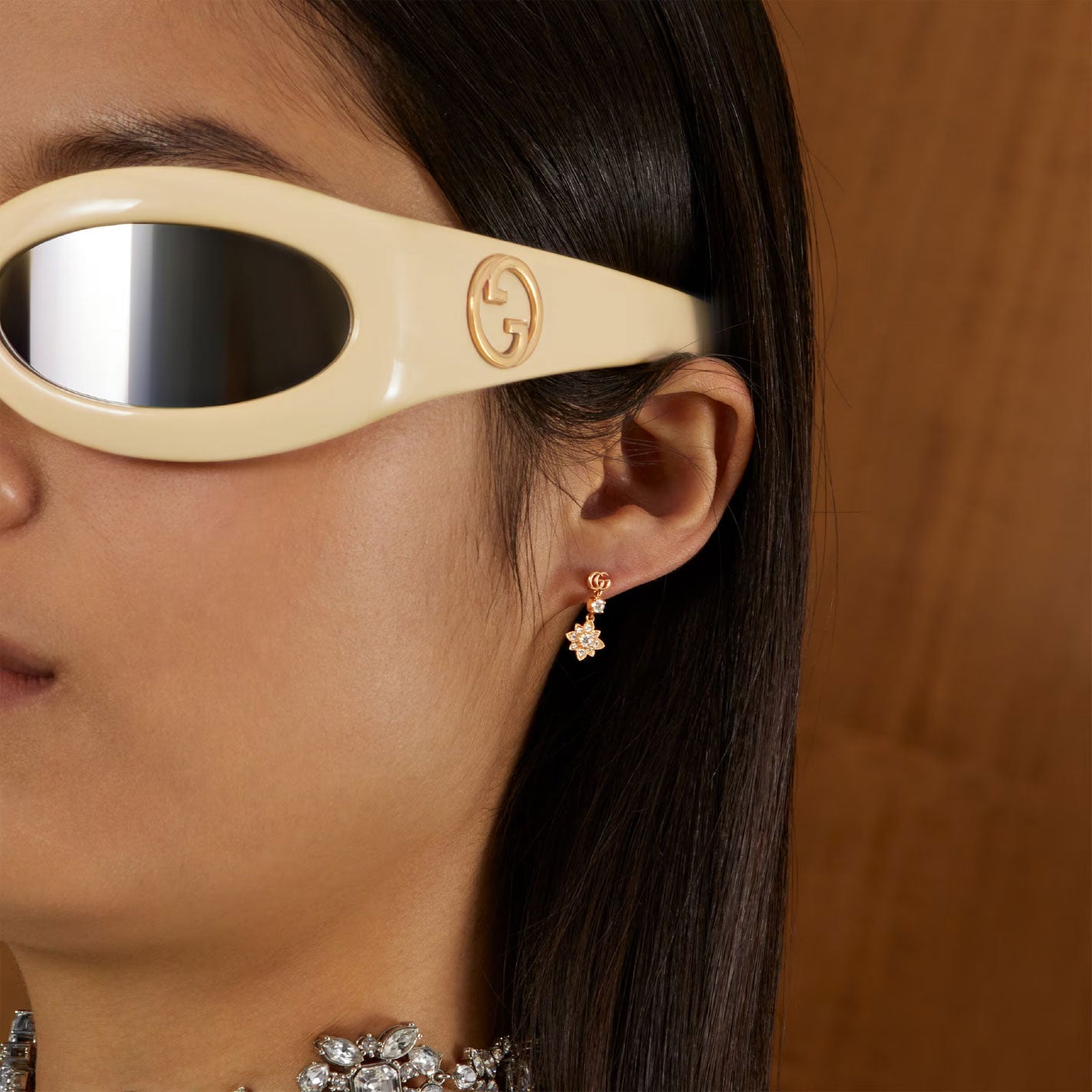 A person dons large, cream-colored sunglasses with a logo and an 18K rose gold diamond necklace. They also wear a pair of small stud earrings from the GUCCI Fine Jewellery collection, specifically the Gucci Flora Double G 18K Rose Gold Diamond Stud Earrings. The background is a warm wooden tone.