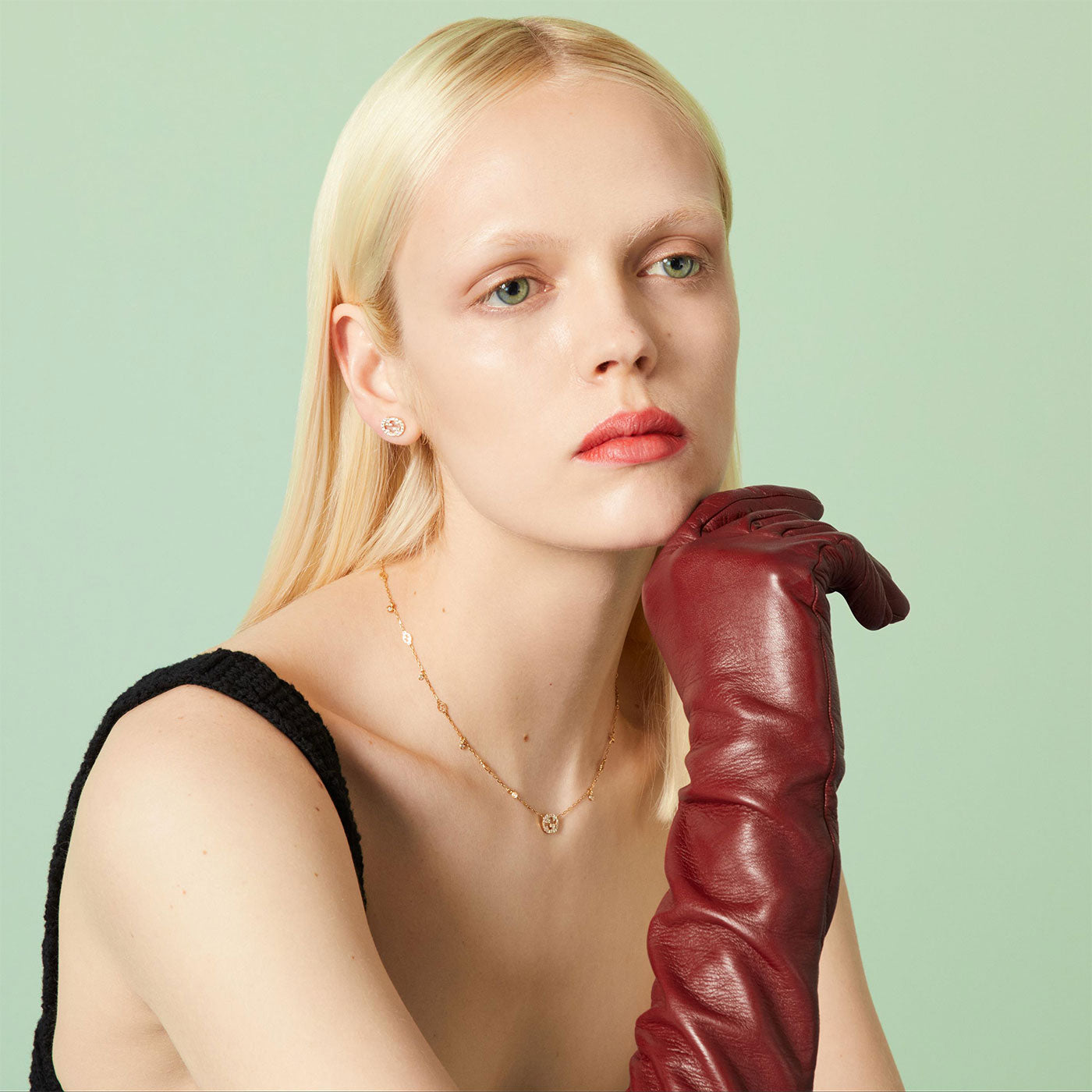 A person with long blond hair wearing a black sleeveless top and a red leather glove on one hand, rests their chin on the gloved hand. They wear the Gucci Interlocking G 18K Yellow Gold Diamond Stud Earrings by GUCCI Fine Jewellery, paired with a simple 18k yellow gold necklace, gazing pensively off-camera. The background is light green.