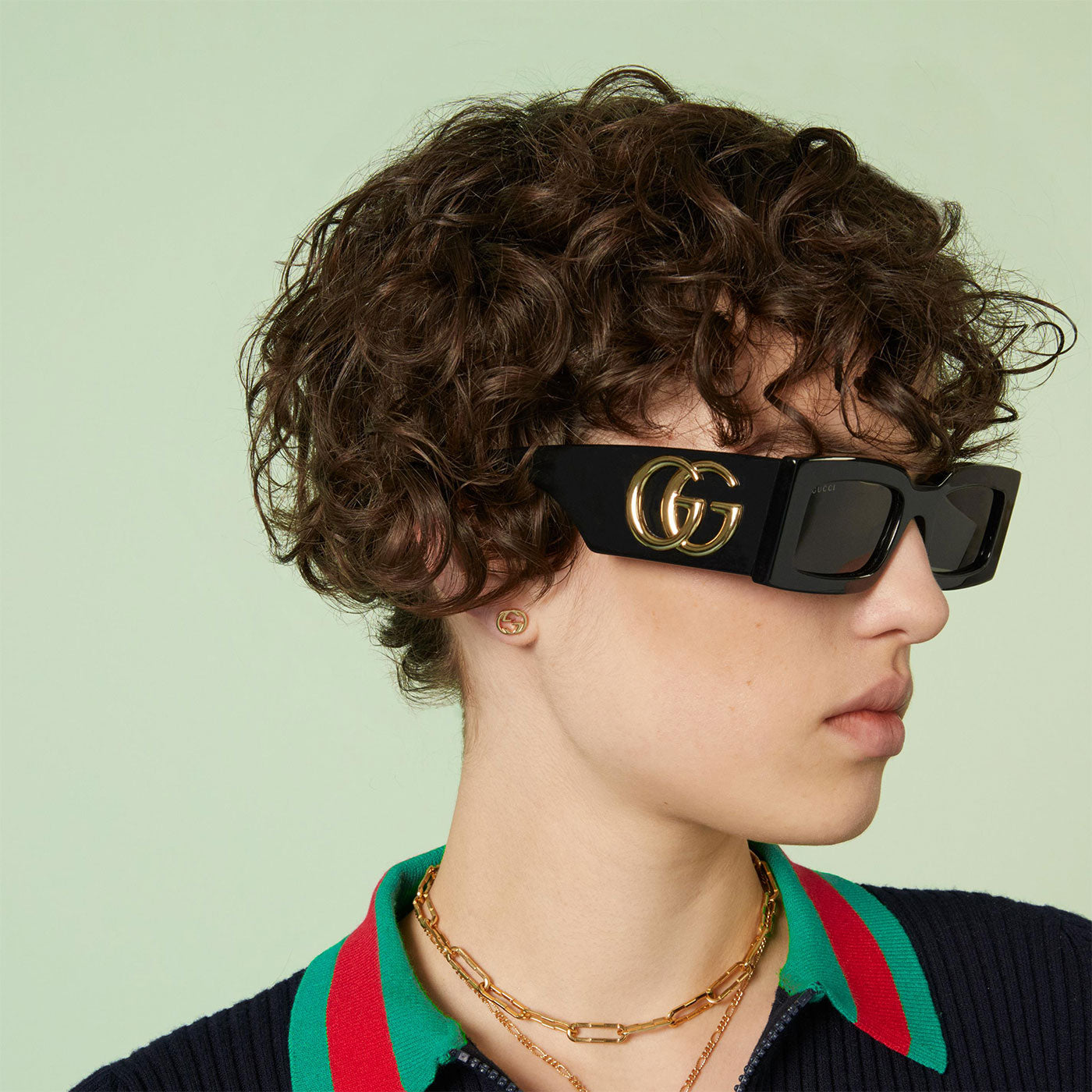 A person with curly hair wears large black sunglasses with gold accents, a striped collared shirt, and a pair of Gucci Interlocking G 18K Yellow Gold Stud Earrings by GUCCI Fine Jewellery against a light green background.