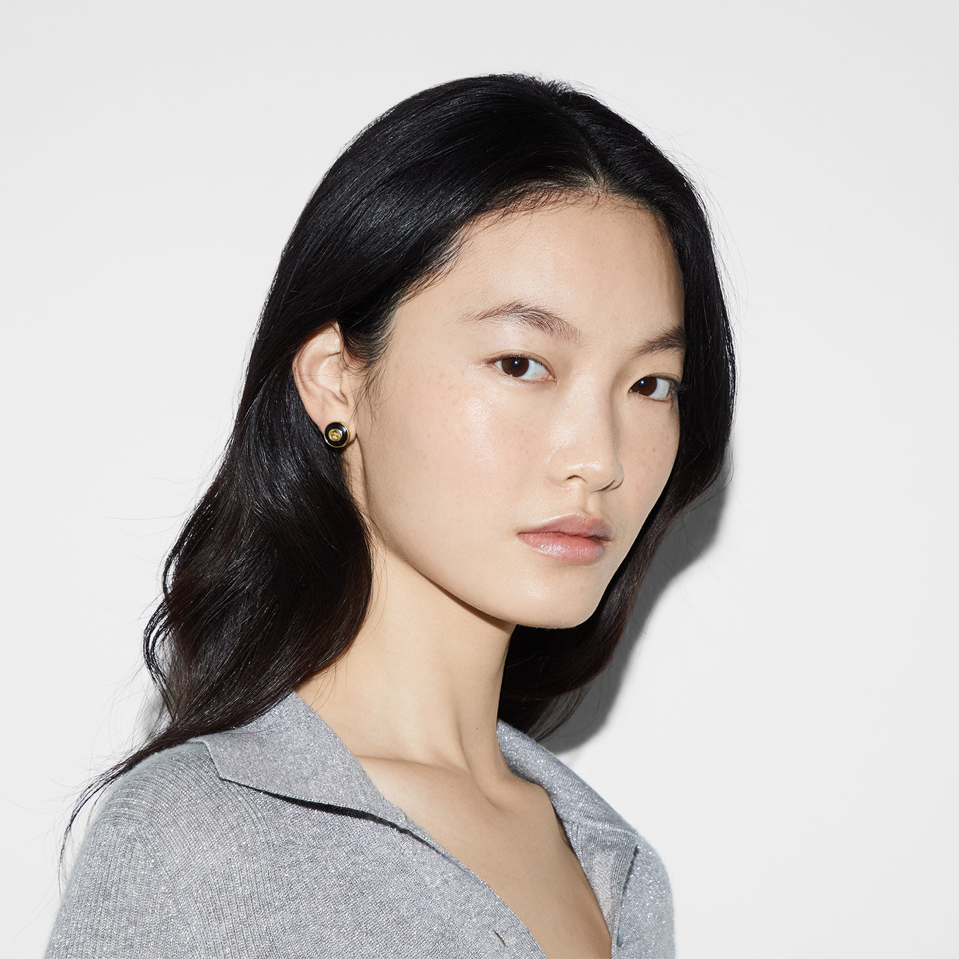 A person with long dark hair is wearing a light gray top and Gucci Interlocking G 18K Yellow Gold and Black Onyx Stud Earrings from GUCCI Fine Jewellery. They are standing against a white background, gazing slightly to the side. The soft, even lighting highlights their facial features, while the subtle sheen of 18k gold adds a touch of elegance.