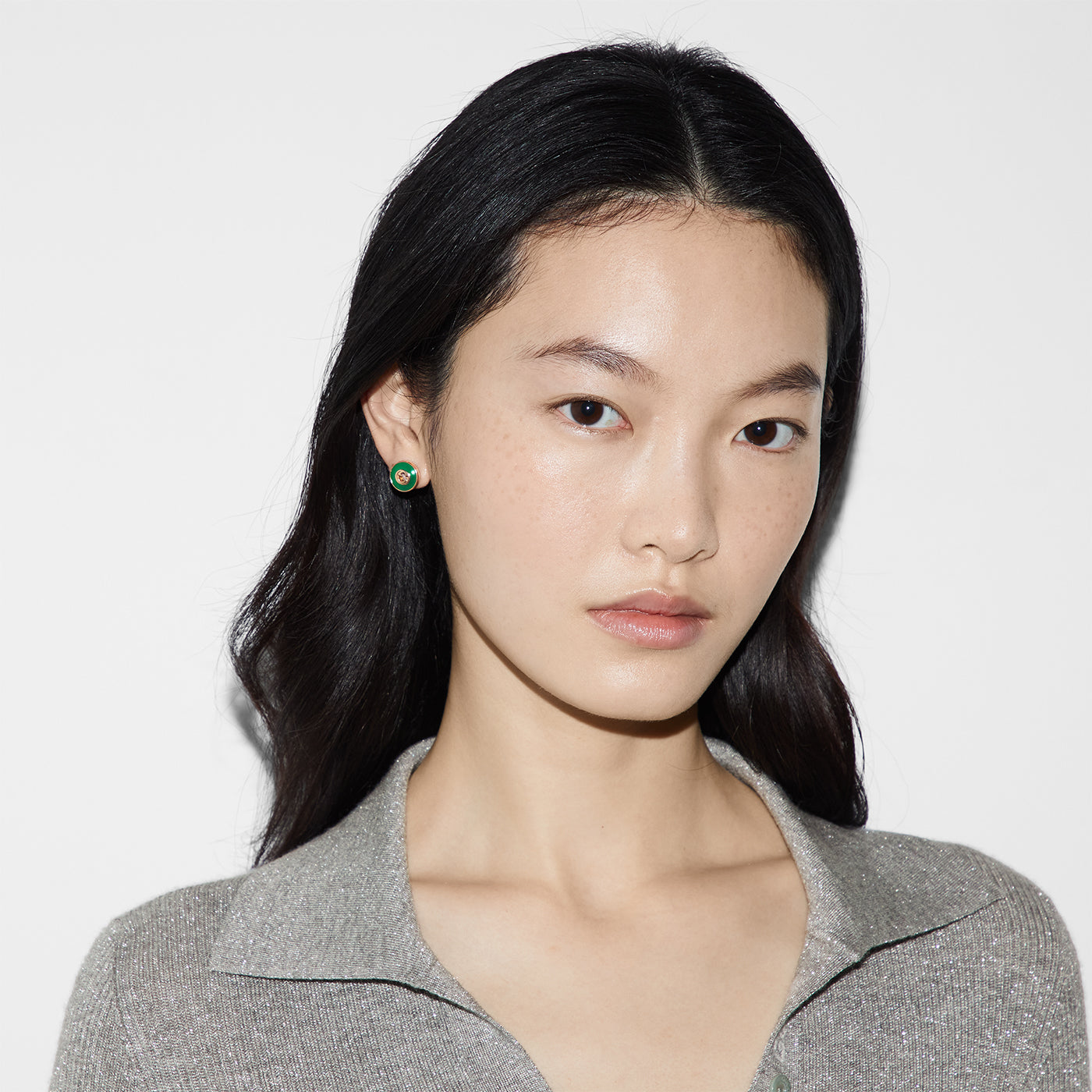 A person with long dark hair and Gucci Interlocking G 18K Rose Gold and Green Agate Stud Earrings from GUCCI Fine Jewellery is wearing a textured grey top, looking directly at the camera against a plain white background.