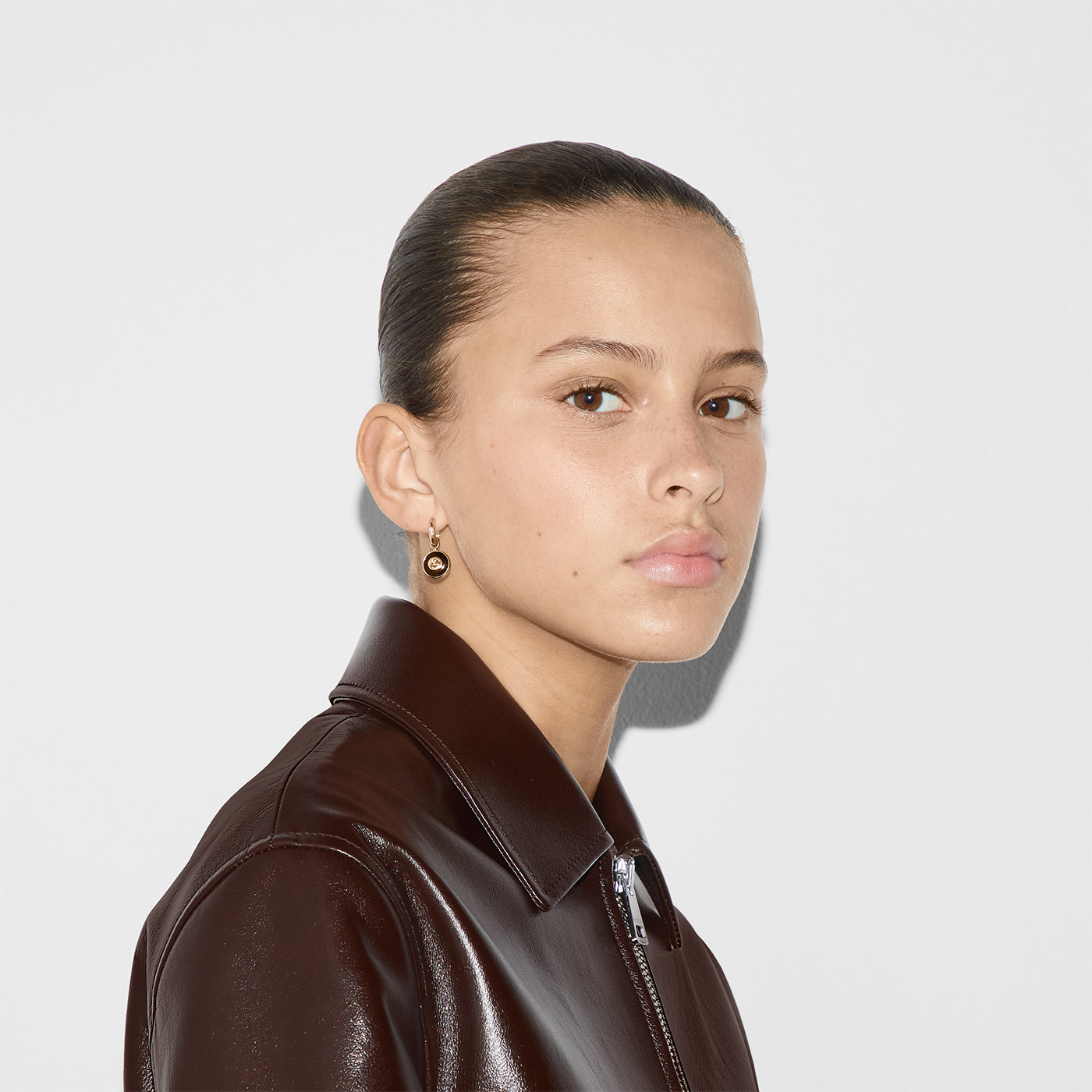 A person with slicked-back hair, wearing a dark leather jacket and a Gucci Interlocking G 18K Yellow Gold and Black Onyx Hoop Earring by GUCCI Fine Jewellery, looks directly at the camera against a plain white background.