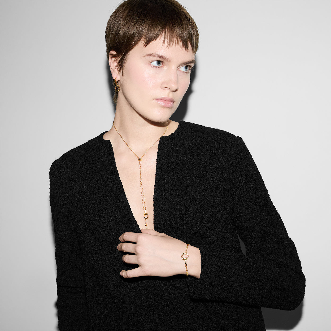 A person with short hair is elegantly dressed in a sleek black outfit, looking to the side. They are adorned with exquisite jewelry, including 18k yellow gold pieces such as a Gucci horsebit bracelet, ring, necklace, and Gucci Horsebit 18K Yellow Gold Earrings from GUCCI Fine Jewellery. The minimalist style is accentuated against a plain white or light gray background.