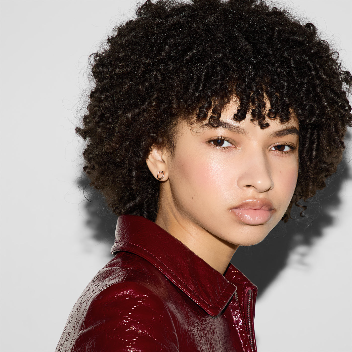 A person with curly hair and a shiny red jacket looks directly at the camera, exuding elegance against a plain background. A touch of luxury gleams from Gucci Horsebit 18K Rose Gold Diamond Stud Earrings by GUCCI Fine Jewellery, accentuating their style effortlessly.