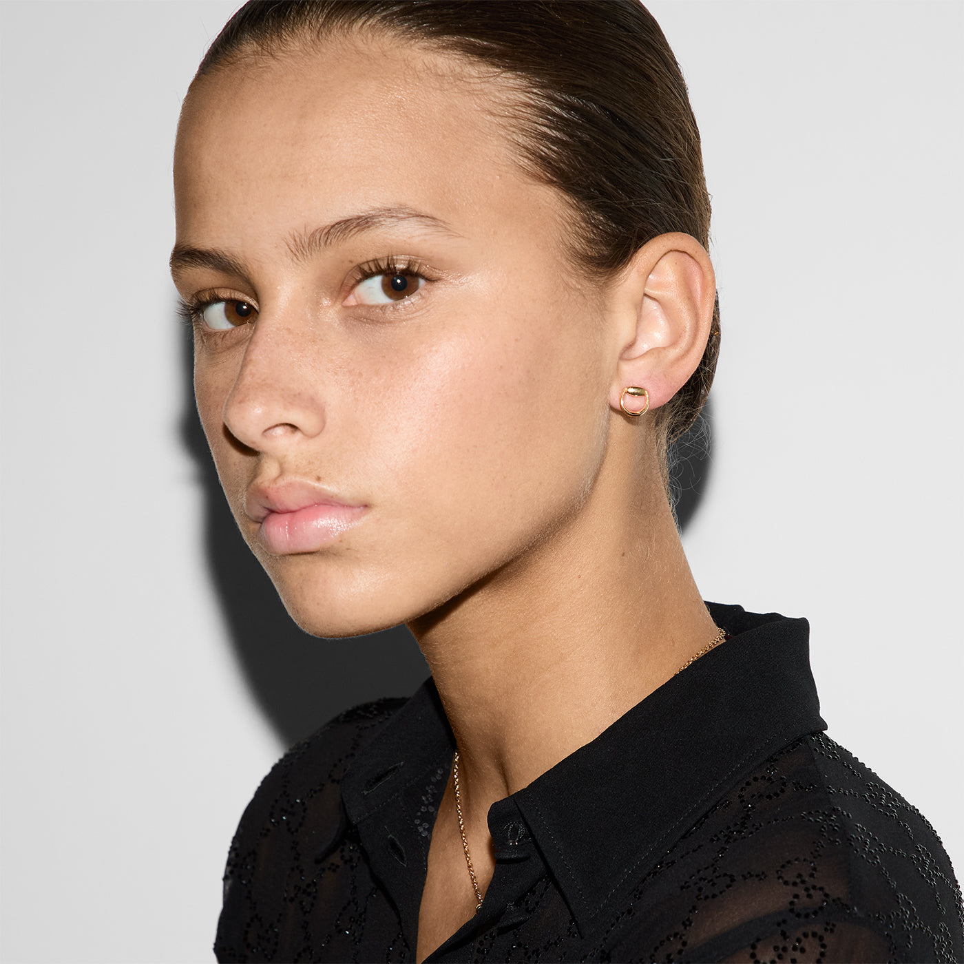 A person with a neutral expression is wearing a black shirt and small, square earrings that catch the light like the Gucci Horsebit 18K Yellow Gold Stud Earrings from GUCCI Fine Jewellery. Their hair is pulled back, and they are facing slightly to the side in front of a plain, light-colored background.