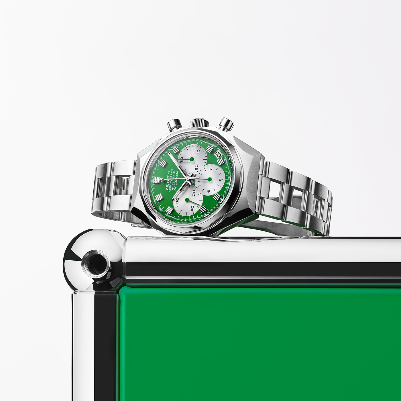 The Zenith Defy Chronograph USM - USM Green, a silver metal wristwatch with a green and white dial, featuring the El Primero 400 calibre, sits on the polished chrome corner of a shiny green and silver box against a white background.