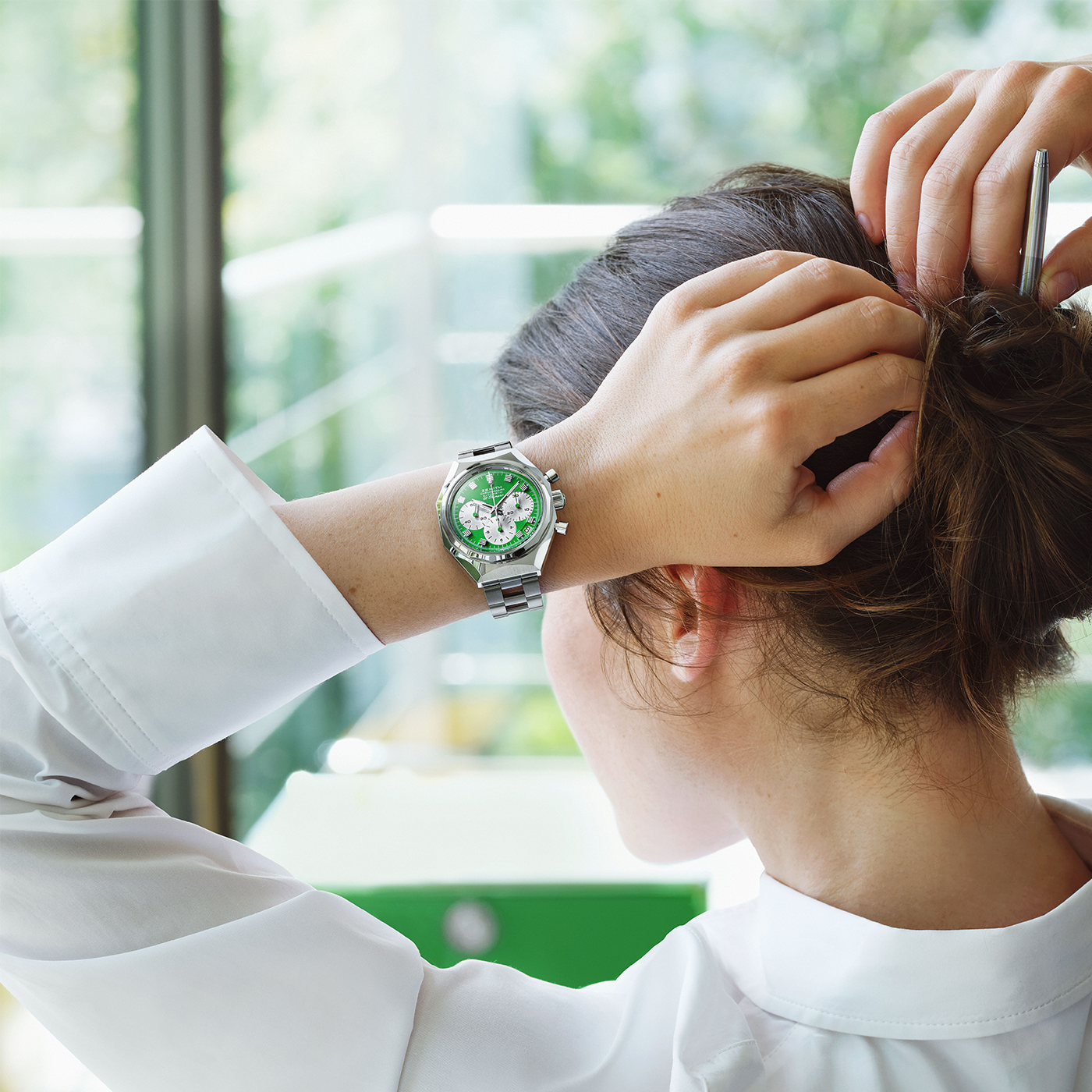 A person with brown hair in a white shirt ties their hair up, revealing a Zenith Defy Chronograph USM - USM Green watch on their left wrist. The background is bright, with blurred greenery and a window.