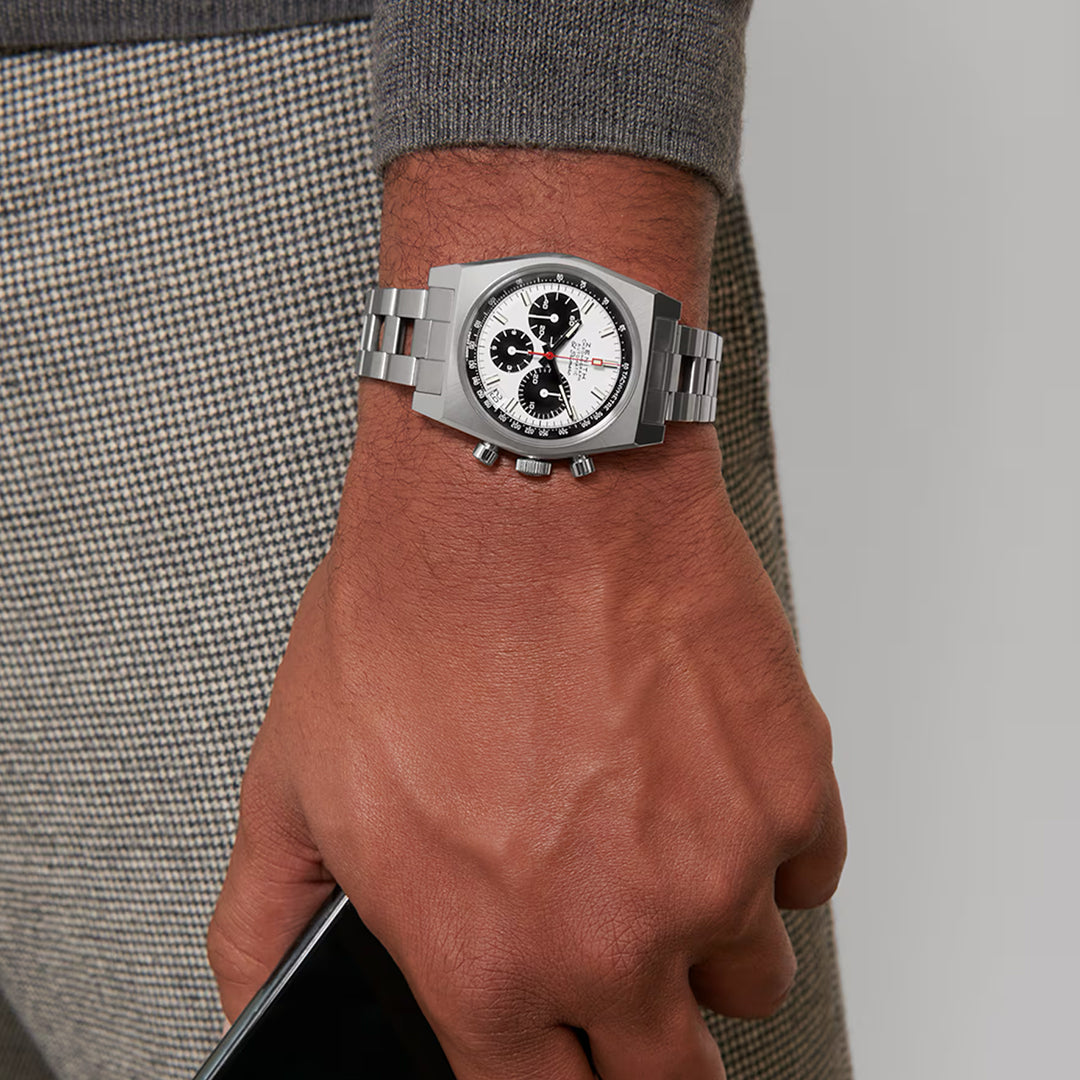 A wrist showcases the ZENITH Chronomaster Revival El Primero 400 37mm, its silver chronograph boasting high-frequency automatic precision. The white face and black dials stand out as the wearer, in a gray sweater and checkered pants, grips a smartphone.
