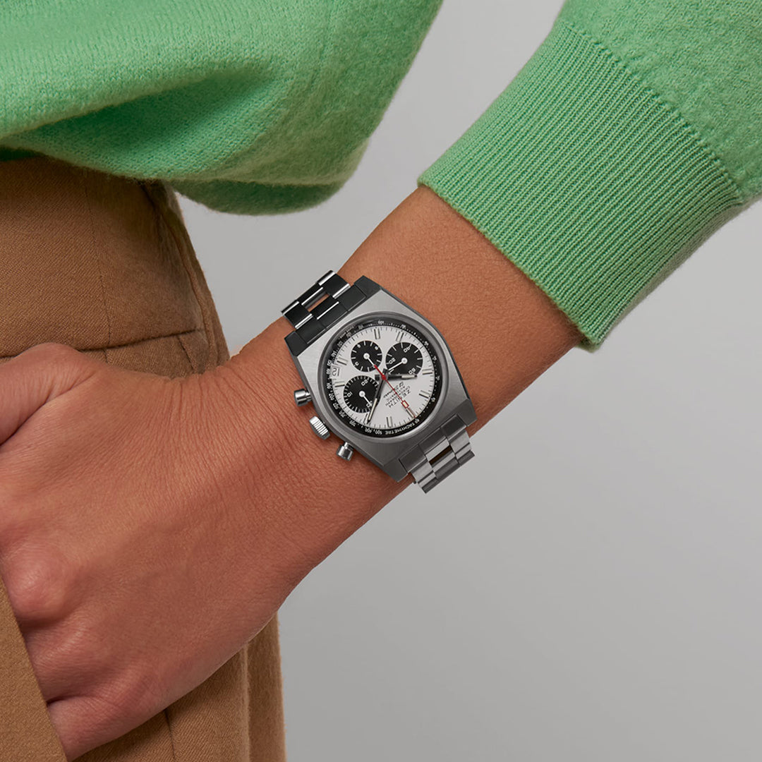 A close-up shows a person wearing the ZENITH Chronomaster Revival El Primero 400 37mm watch. The silver Zenith timepiece with a white and black dial complements their green sweater and brown pants, as their hand rests confidently on their hip.