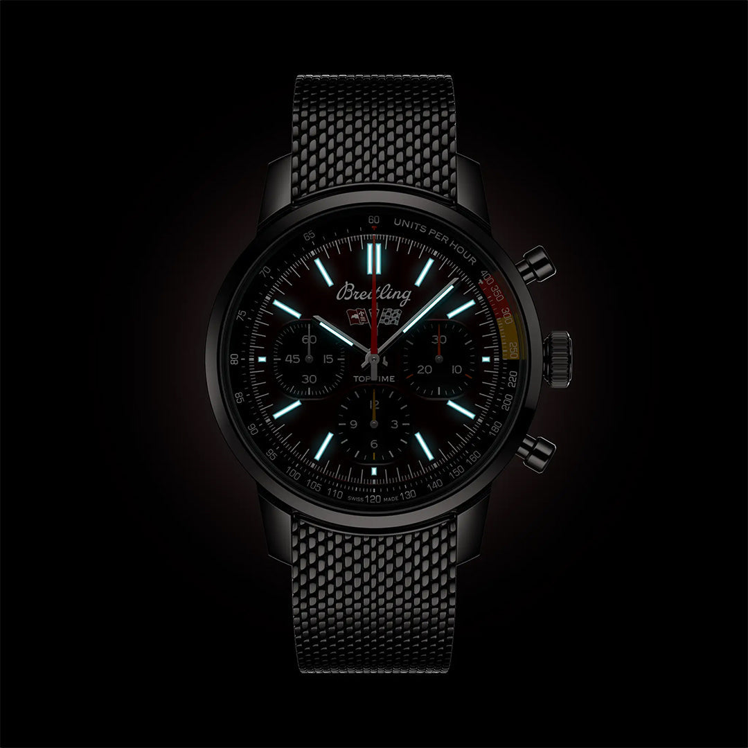 The Breitling Top Time B01 41 Chevrolet Corvette is a black chronograph wristwatch with a mesh strap, blue-lit hour markers, three subdials, and a tachymeter—classic car-inspired and set against a dark background.