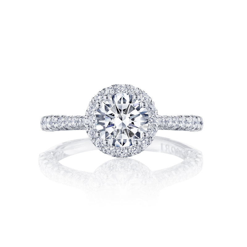 The Tacori Petite Crescent 18K White Gold Round Brilliant Semi Mount Engagement Ring features a central large round diamond encircled by a halo of smaller diamonds. Its band is embellished with pave-set diamonds and boasts a cathedral foundation, all showcased vertically on a reflective white surface to highlight the brilliance of Tacori gemologists' craftsmanship.