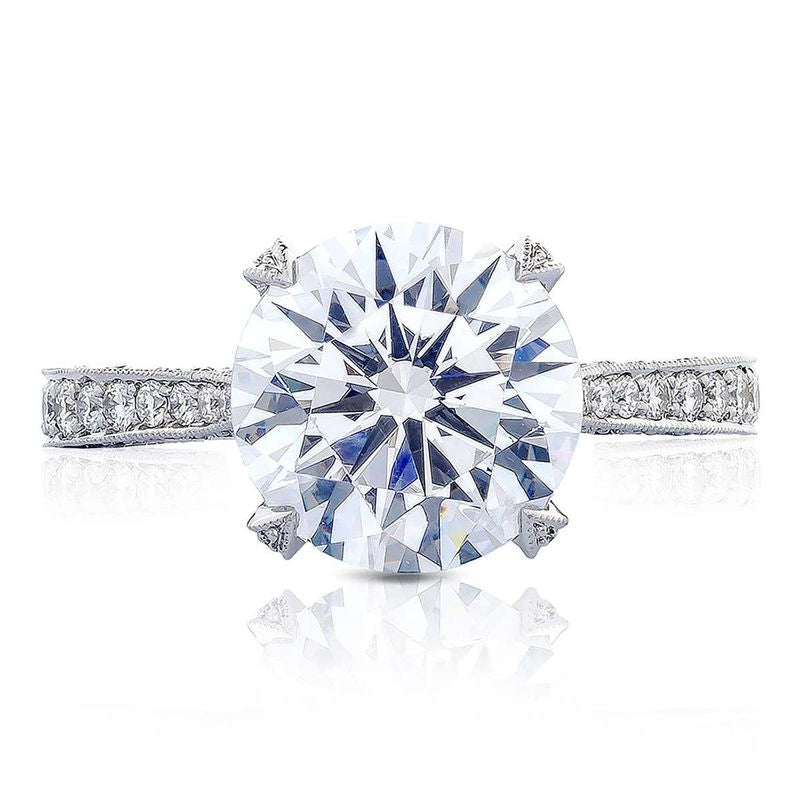 A close-up of the Tacori Classic Crescent RoyalT Round Brilliant Solitaire Semi Mount with Diamond Band Engagement Ring highlights a prominent round-brilliant diamond at its center, elegantly set on a band adorned with smaller diamonds. The design includes four prongs securing the central diamond, meticulously crafted by TACORI gemologists.